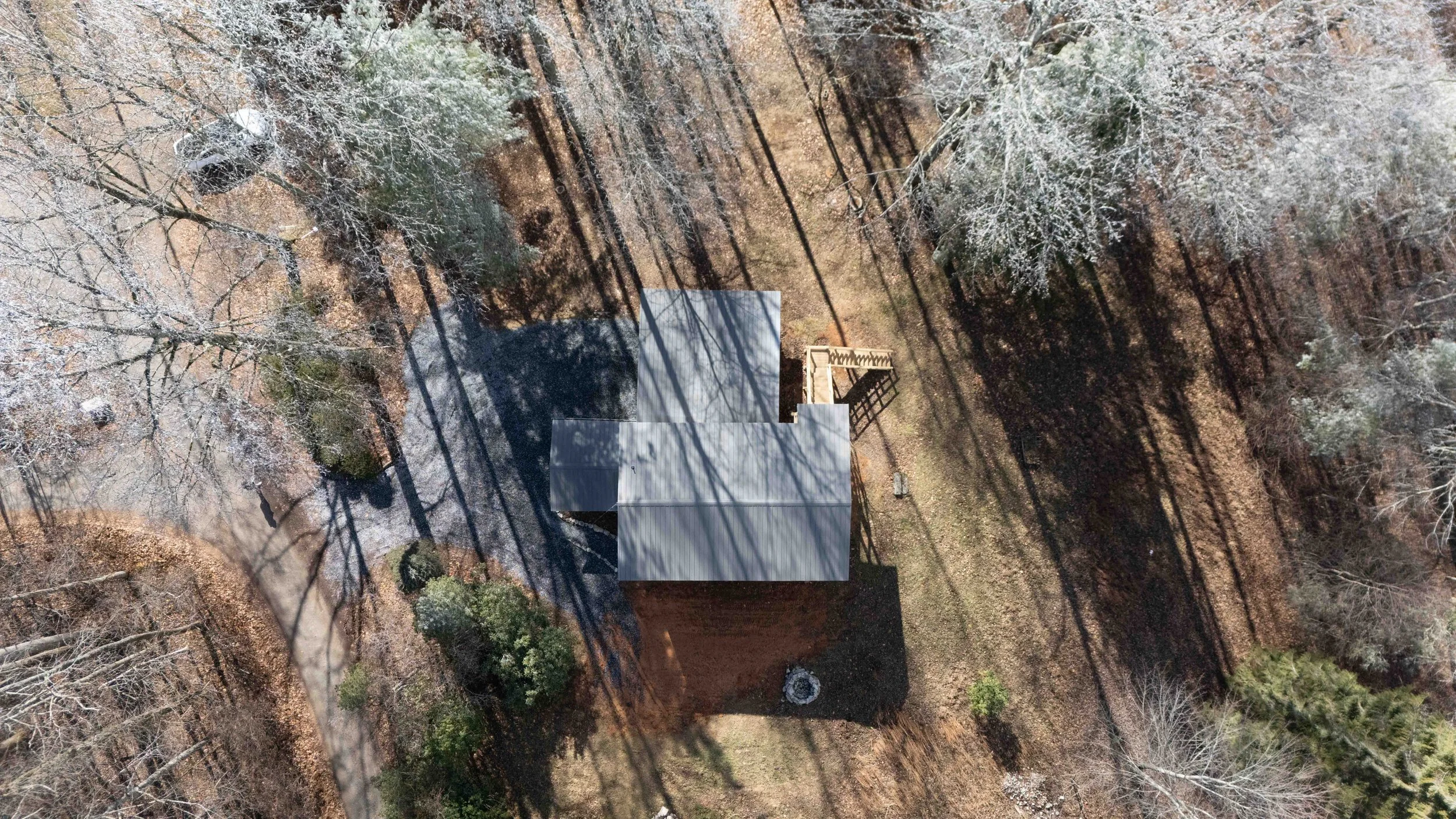 Aerial view of a house with a grey roof surrounded by leafless trees and a wooden deck, casting long shadows on the ground.