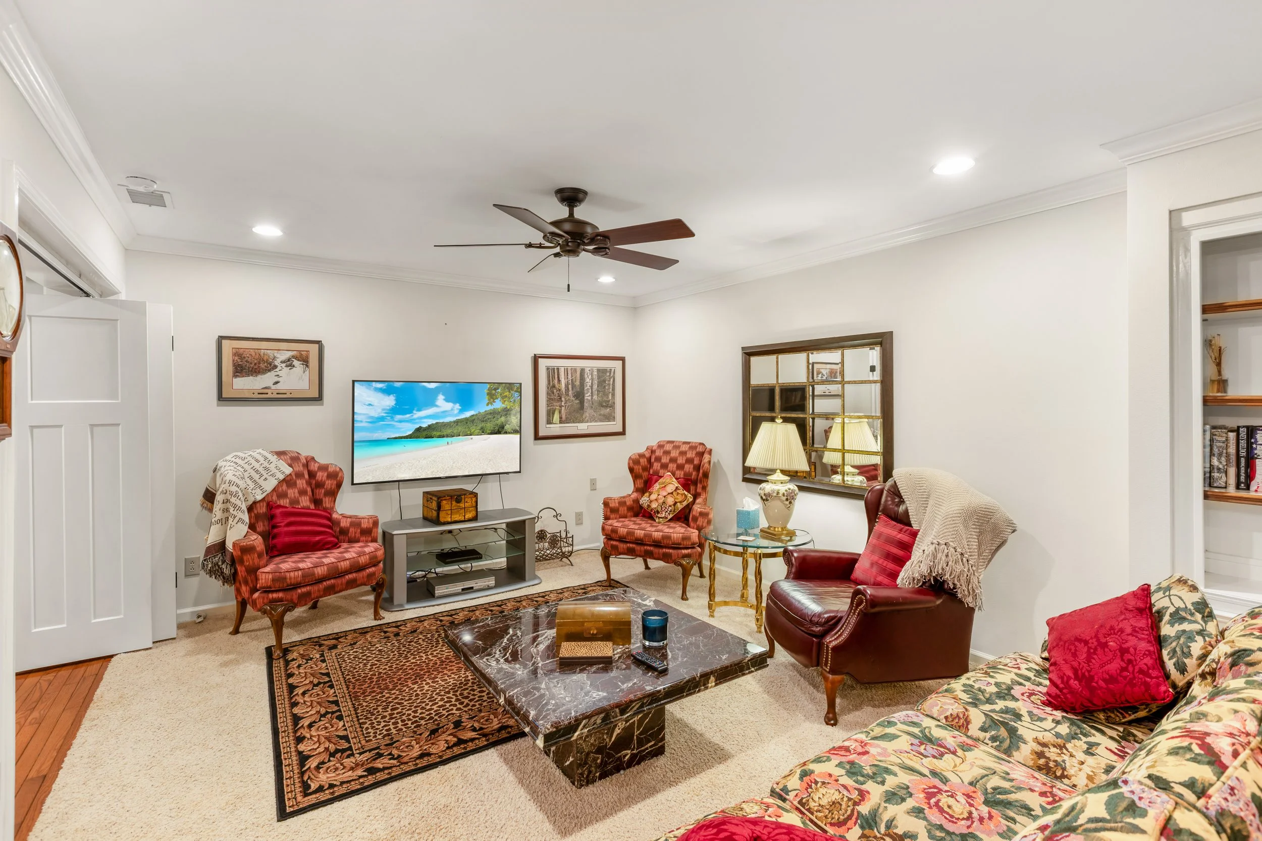 Living room with white walls, beige carpet, a marble coffee table, patterned sofa with red pillows, leather armchair with a cream throw, two red checkered armchairs, a TV on a gray stand, and various framed pictures on the walls. There are lamps, a w
