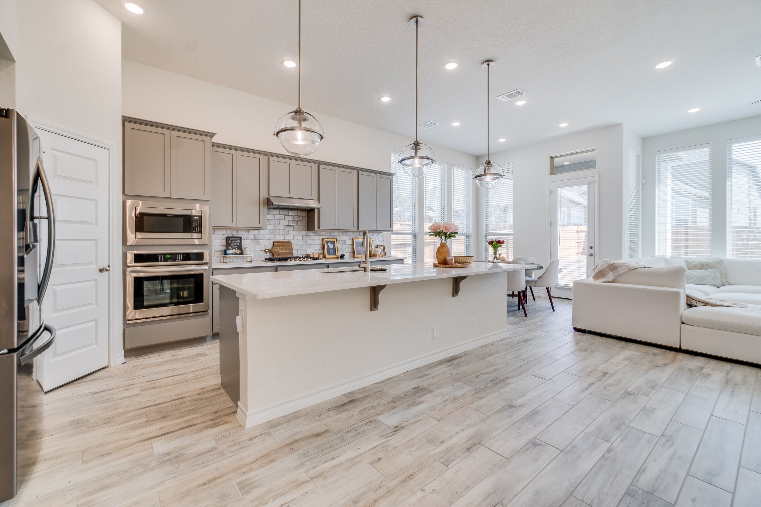 Open-concept kitchen and living area with light gray cabinets, white brick backsplash, stainless steel appliances, a large island with a white countertop, and a seating area with white sofa and dining table near windows with blinds.