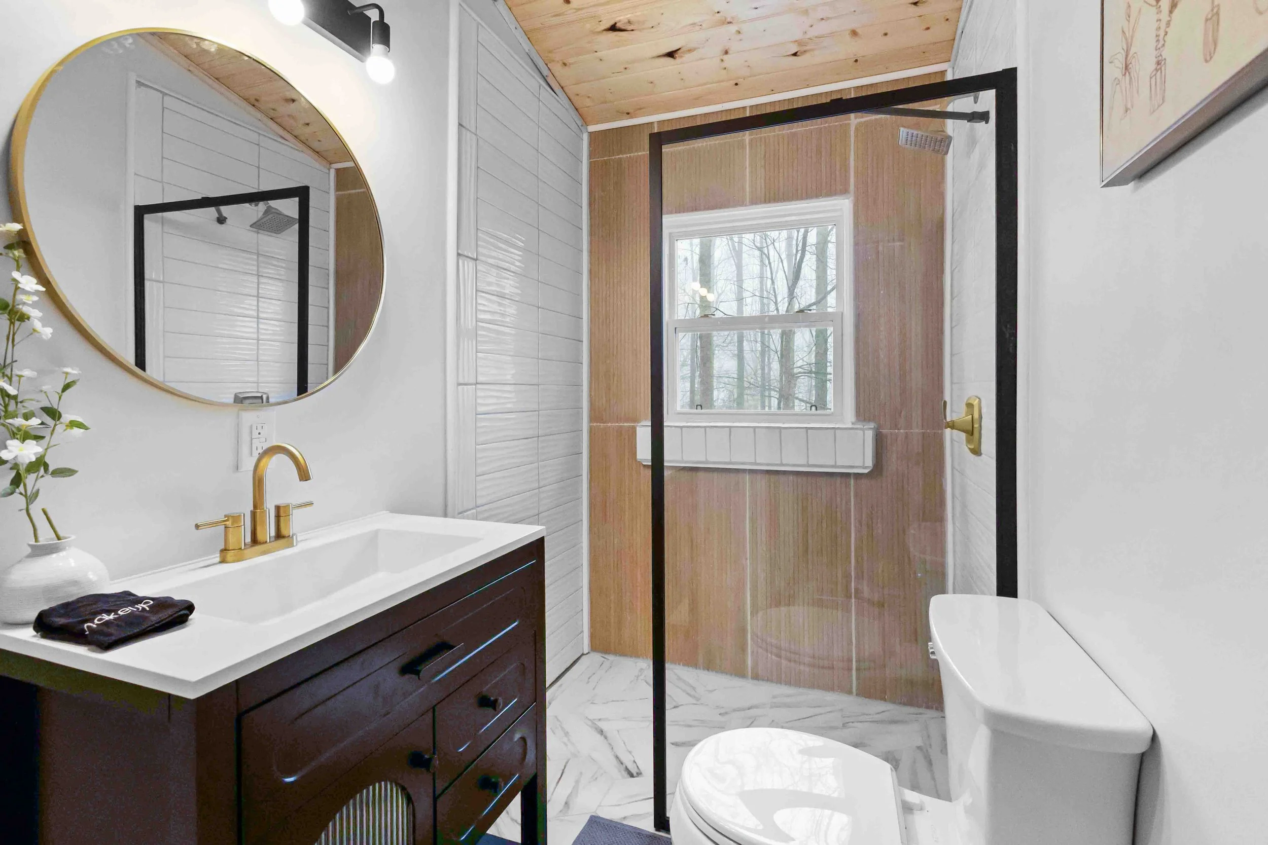 A modern bathroom with a white vanity, gold fixtures, round mirror, black trim glass shower enclosure, marble floors, and a wooden ceiling. There is a window in the shower area with trees visible outside.