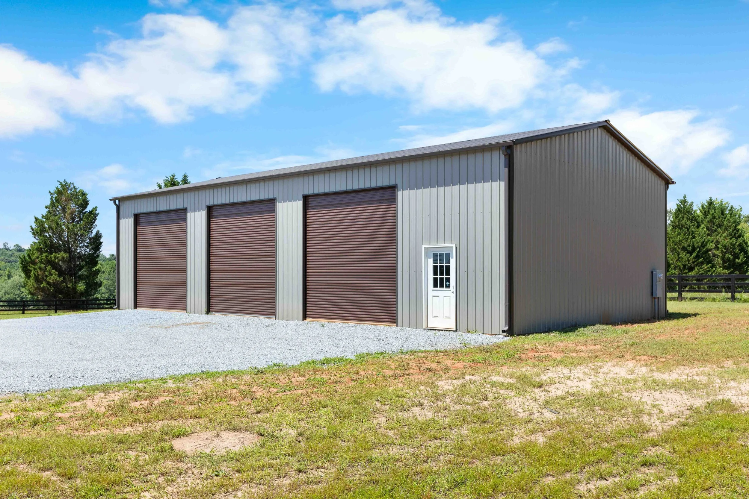 Large metal storage building with three brown roll-up doors and a small white door on the side, situated on a gravel driveway with grass in front, surrounded by trees under a partly cloudy sky.
