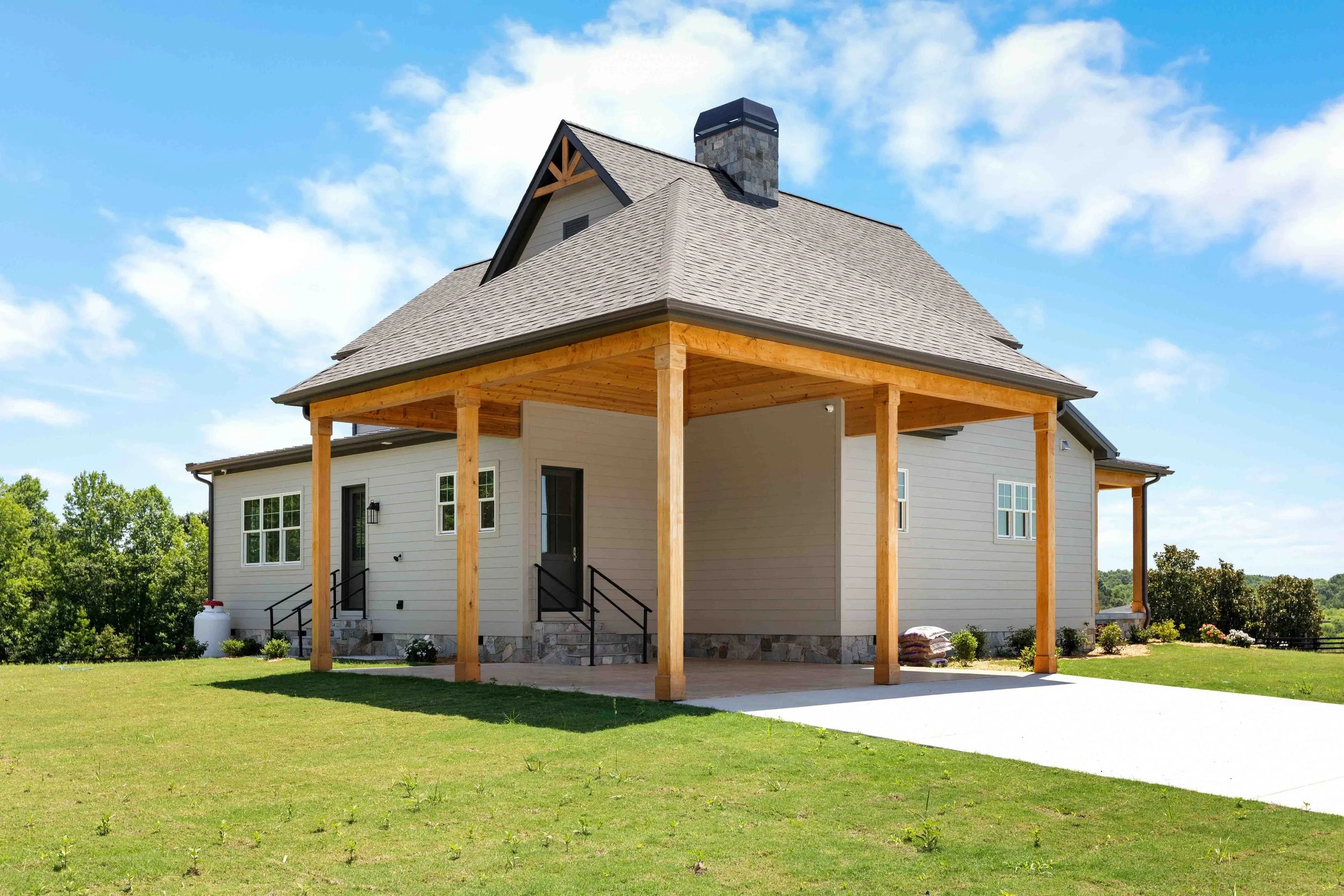 Newly built house with a wraparound porch, black railing, and a large front yard under a partly cloudy sky.