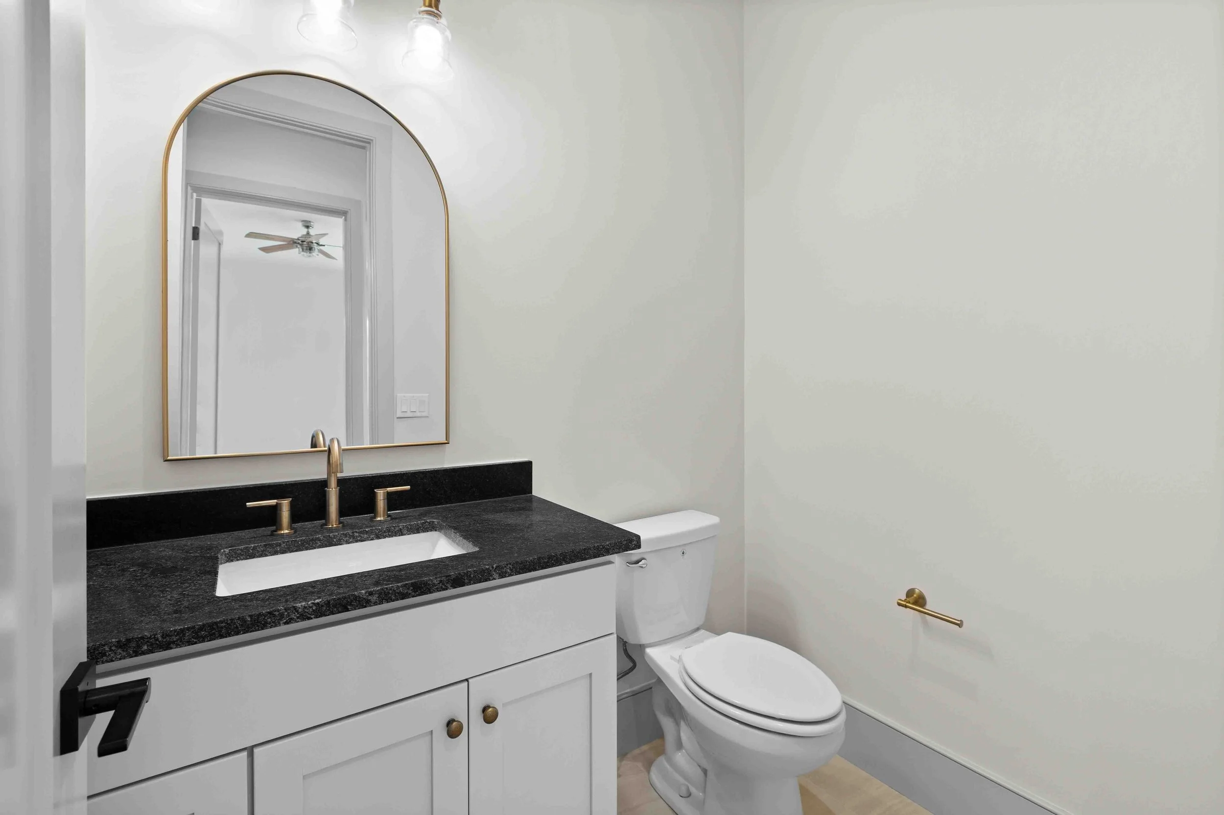 Bathroom with white walls, black countertop sink with gold fixtures, a mirror with a gold frame, and a white toilet. There is a towel bar on the wall and a ceiling light with a visible bulb.