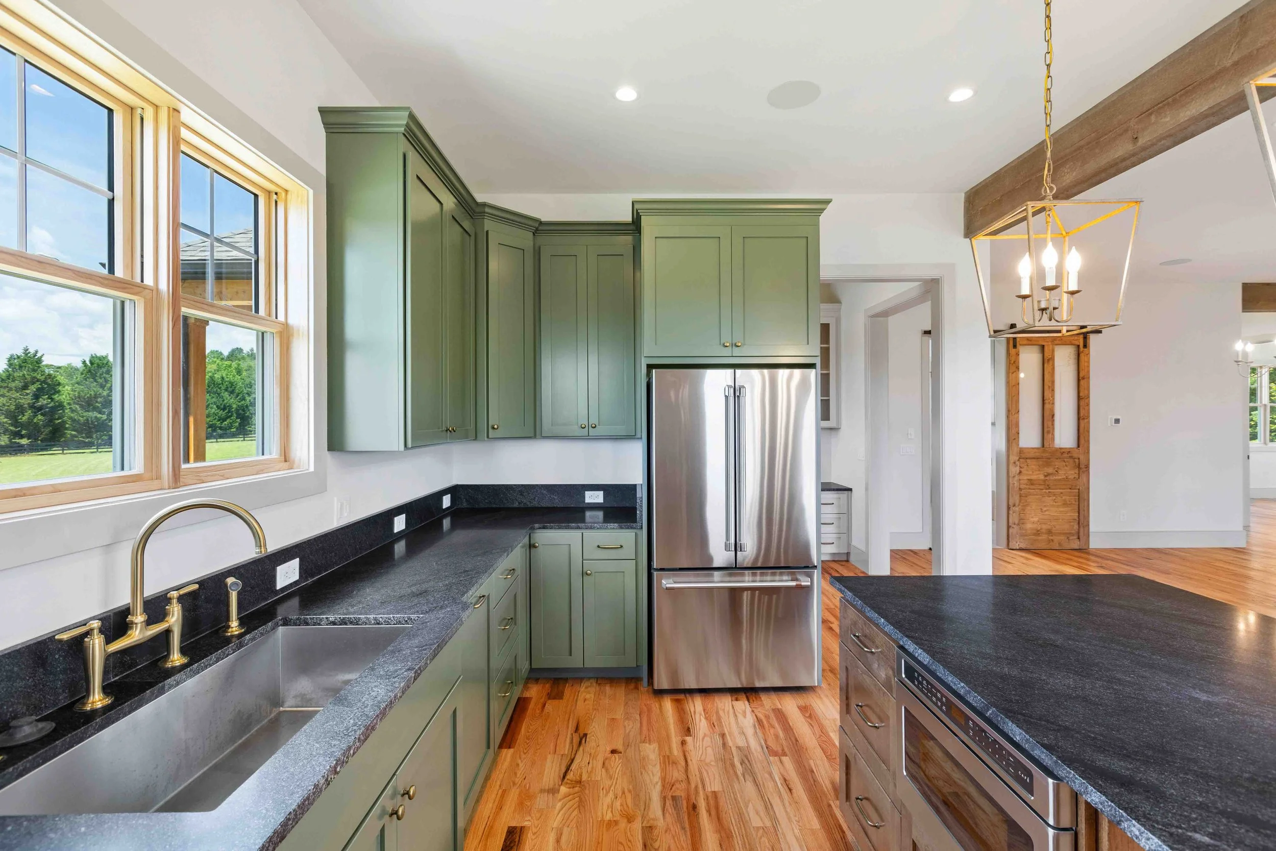 Modern kitchen with green cabinets, black countertops, stainless steel refrigerator, hardwood floors, and large windows overlooking a green outdoor landscape.
