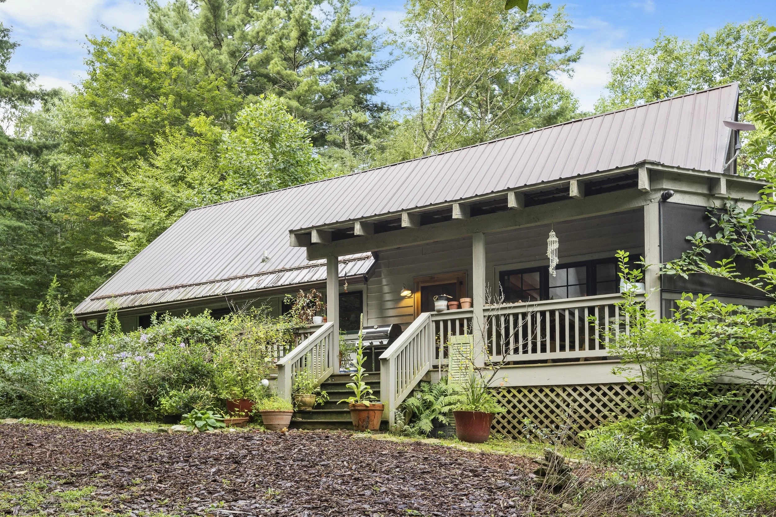 A house with a gray metal roof surrounded by lush green trees and plants, with a small porch, potted plants, and decorative hanging ornament.