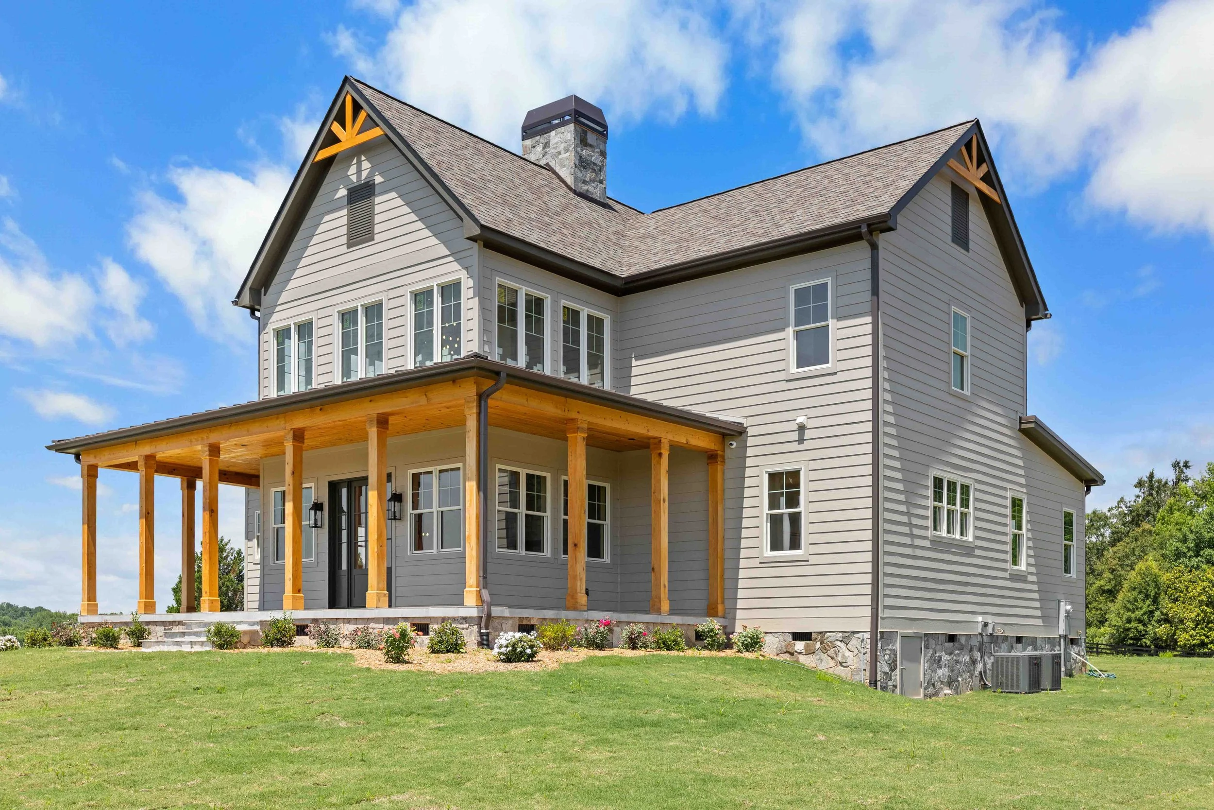 A two-story house with beige siding, multiple windows, a large chimney, and a covered porch with wooden support beams, set on a grassy lawn under a partly cloudy blue sky.