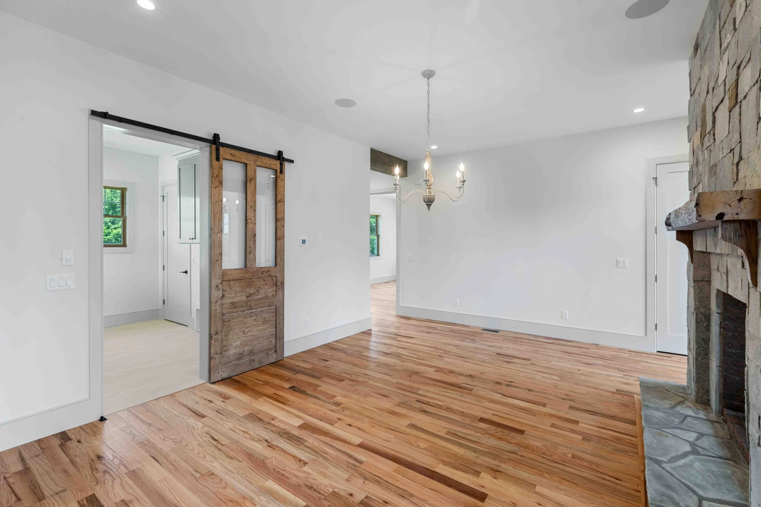 Empty living room with hardwood floors, white walls, a stone fireplace, a chandelier, and a sliding barn door.