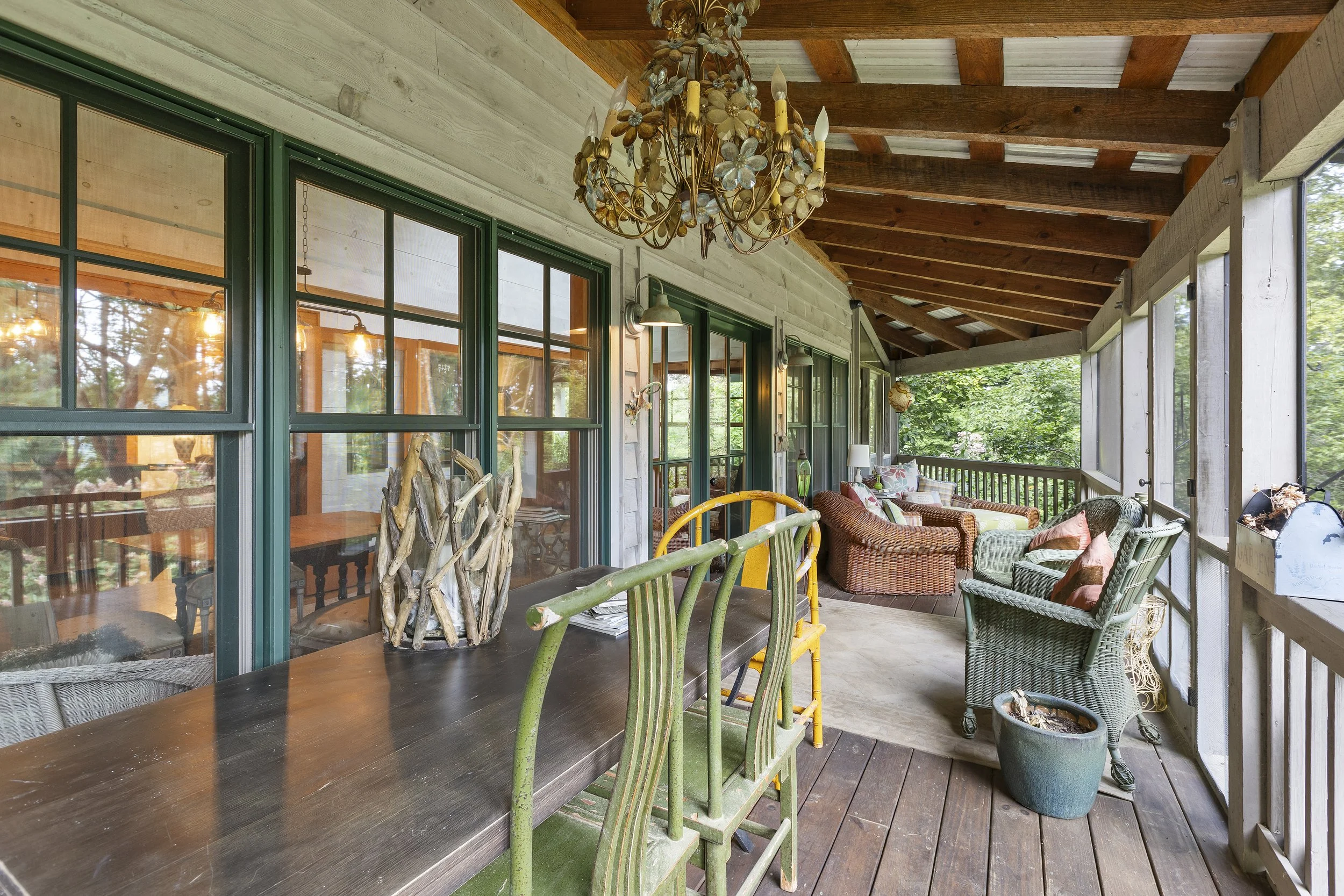A covered porch with wooden flooring, green-framed windows, a chandelier, outdoor seating including wicker chairs and sofas, and decorative plants.