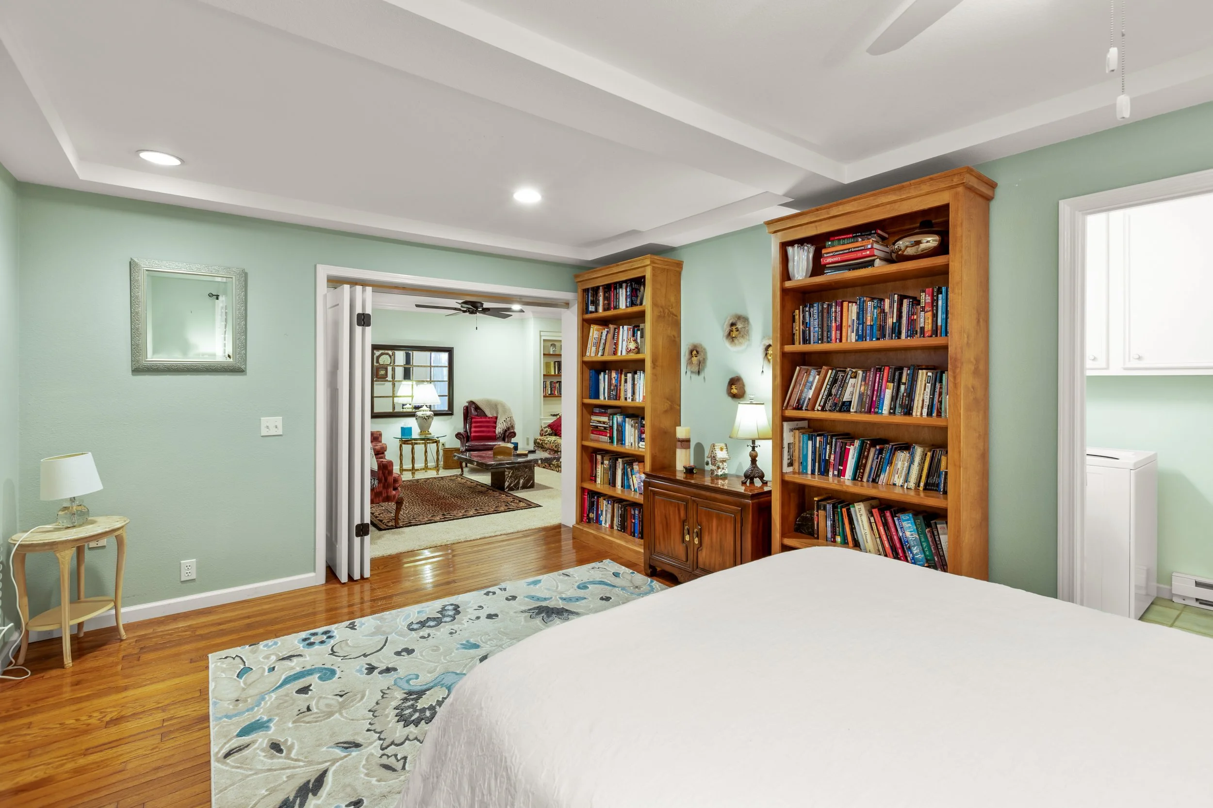 Bedroom with green walls, wooden bookshelves filled with books, a nightstand with a lamp, part of a bed with white bedding, and an open doorway leading to a living room with additional seating and a rug.