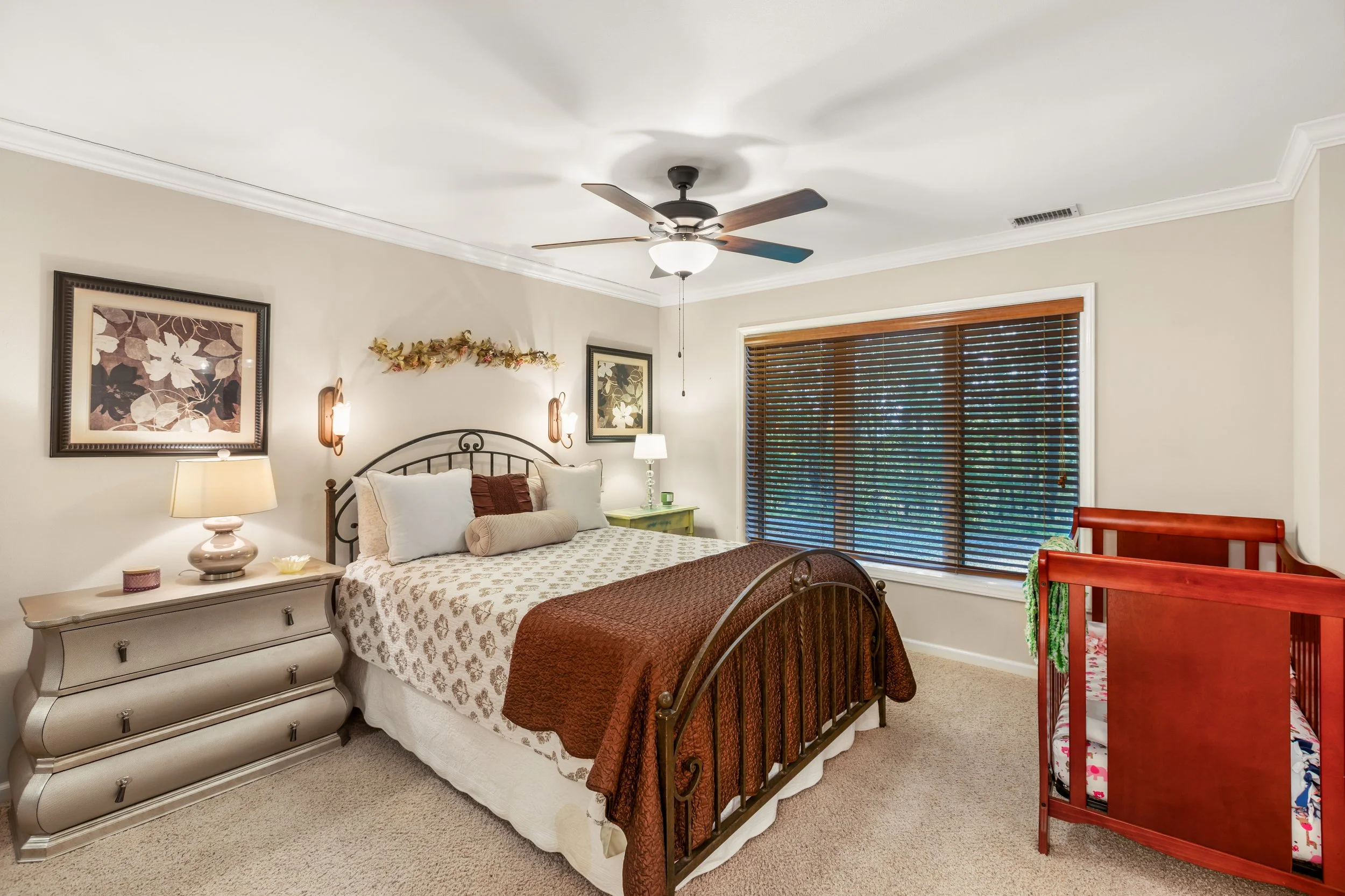 A bedroom with cream-colored walls, a queen-sized bed with a black metal headboard, various pillows, and a brown quilt. To the left of the bed is a white dresser with a beige lamp, a small container, and a decorative item. On the wall above the bed a