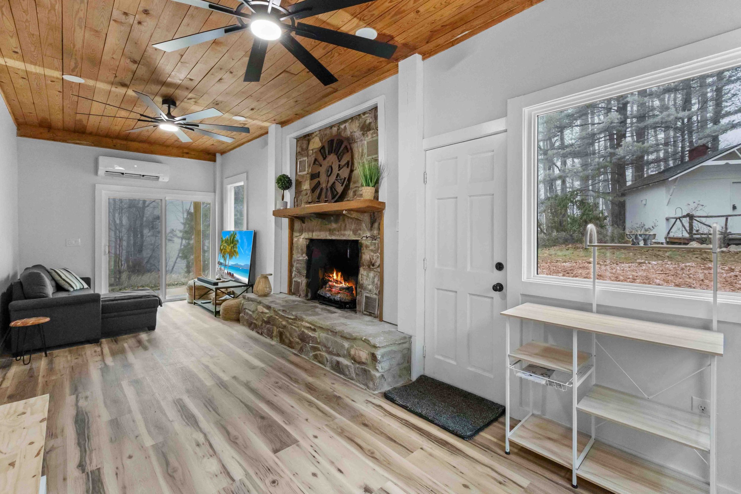 Living room with a stone fireplace, a ceiling fan, light wood flooring, a gray sofa, a TV displaying a beach scene, and large windows showing an outdoor wooded yard.