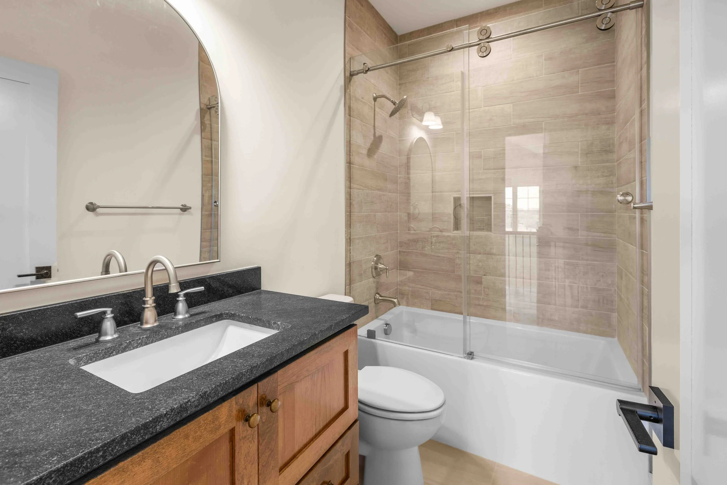 Bathroom with a black granite countertop vanity, wooden cabinet, large mirror, white sink, silver faucet, glass shower with beige tiles, bathtub, and white toilet.
