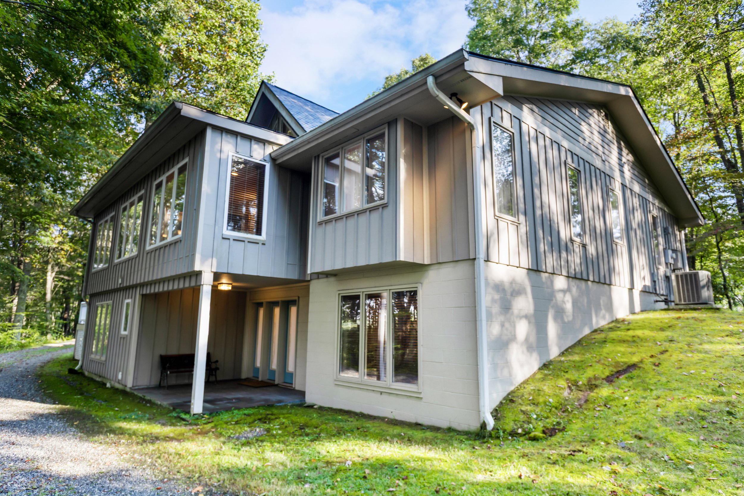 A two-story house with gray siding, multiple windows, and a sloped roof, situated in a wooded area with green grass and trees.