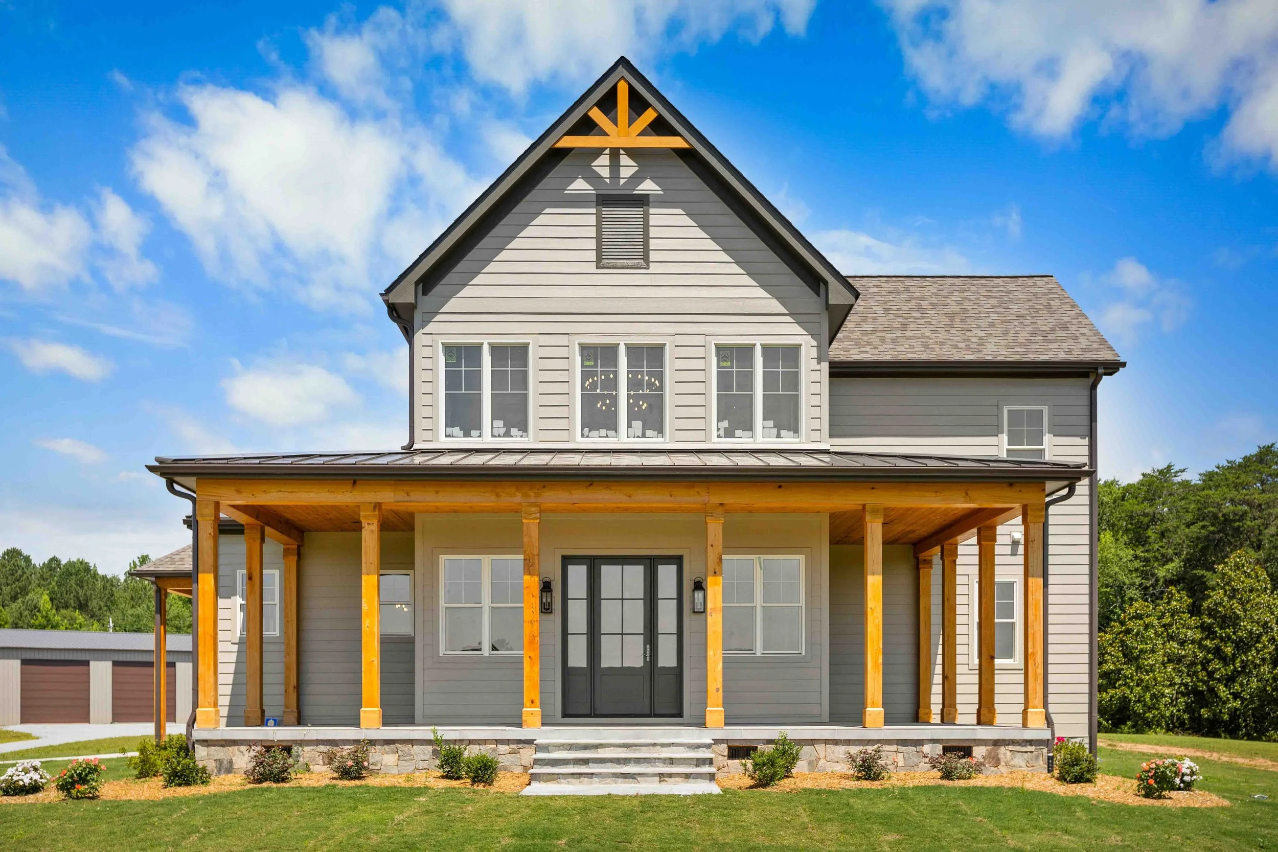 Front view of a modern house with a gray exterior, front porch with wooden columns, and a well-maintained lawn under a partly cloudy sky.