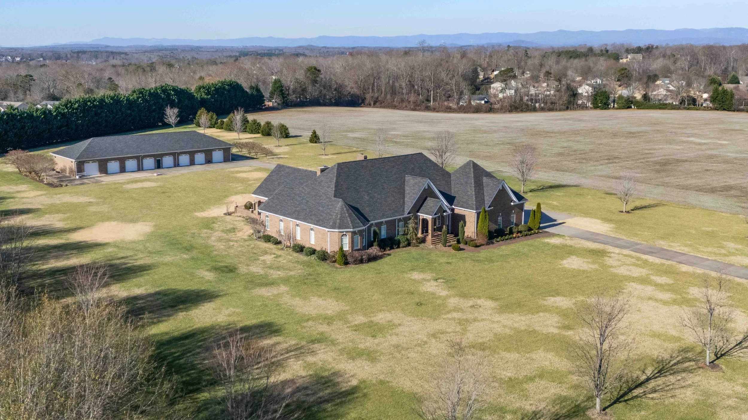 Aerial view of a large brick house with a dark gray roof, surrounded by a spacious lawn with a few leafless trees. A paved driveway leads to the house and a detached garage. In the background, there is an open field, a row of evergreen trees, and dis