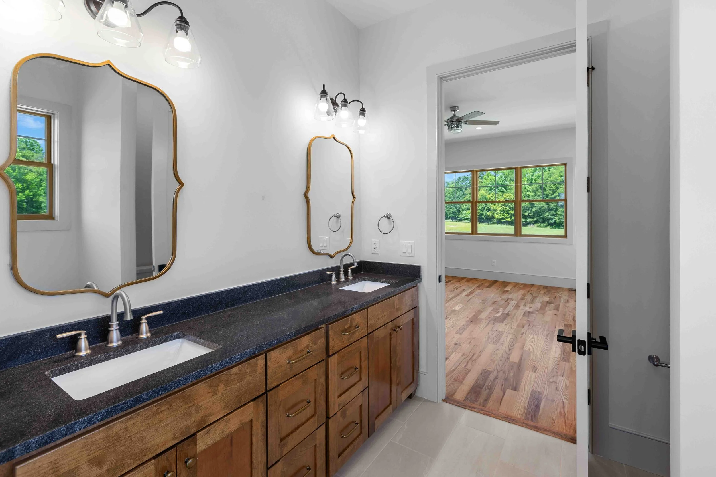Bathroom with dual sinks, wooden cabinets, black countertop, oval mirrors with gold edges, and three wall-mounted light fixtures. Through the doorway, a room with large windows, hardwood flooring, and a ceiling fan is visible.