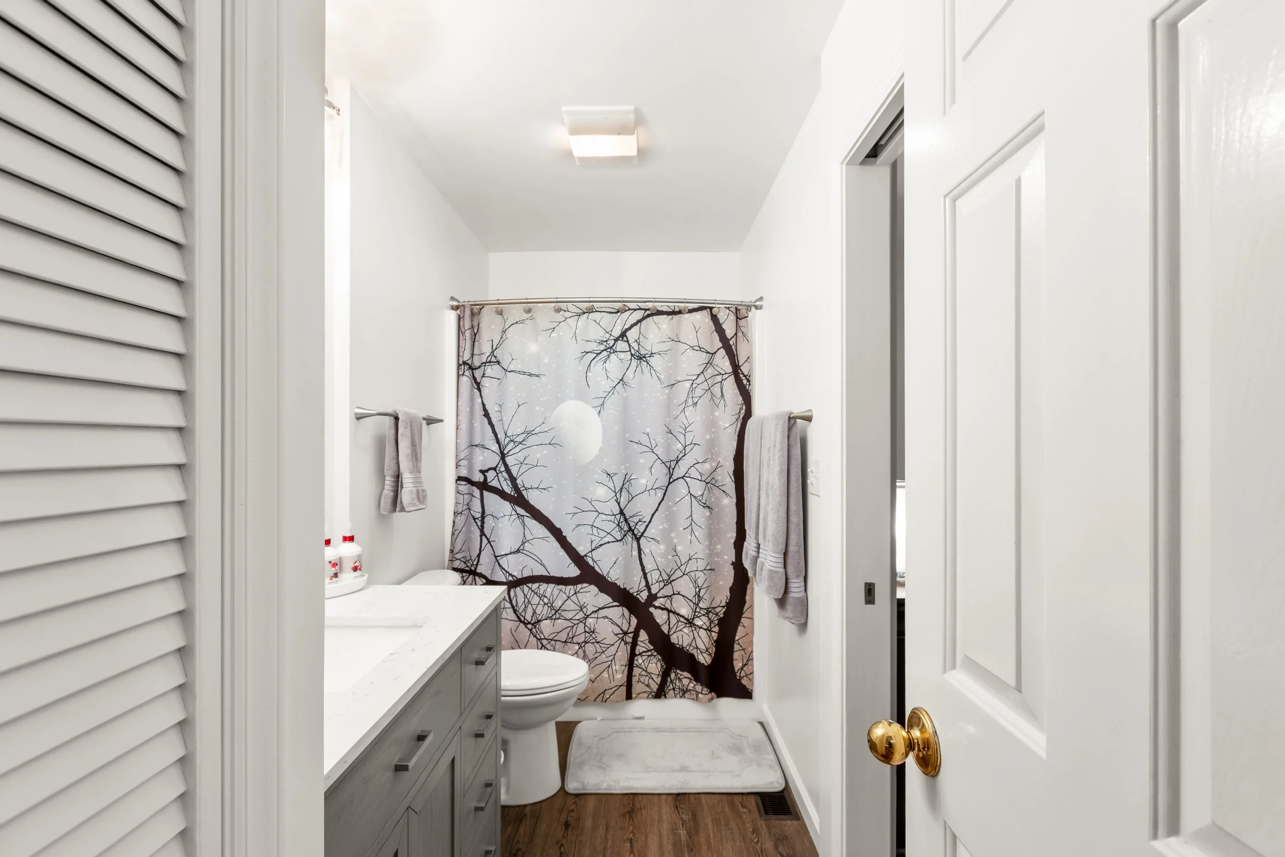 A small bathroom with white walls and a white ceiling. There is a shower with a curtain showing a scene of leafless tree branches and a full moon. The vanity counter supports some small bottles, and two gray towels hang on a towel bar. There is a whi