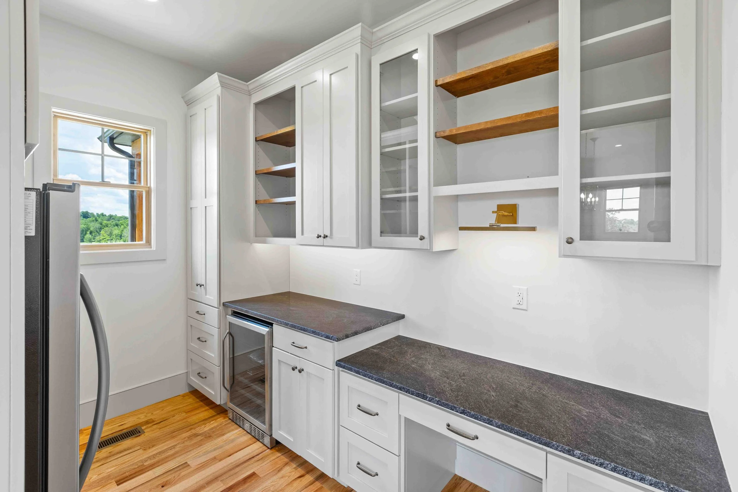 Kitchen with white cabinets, dark countertops, a window with a view of greenery, and hardwood floors.