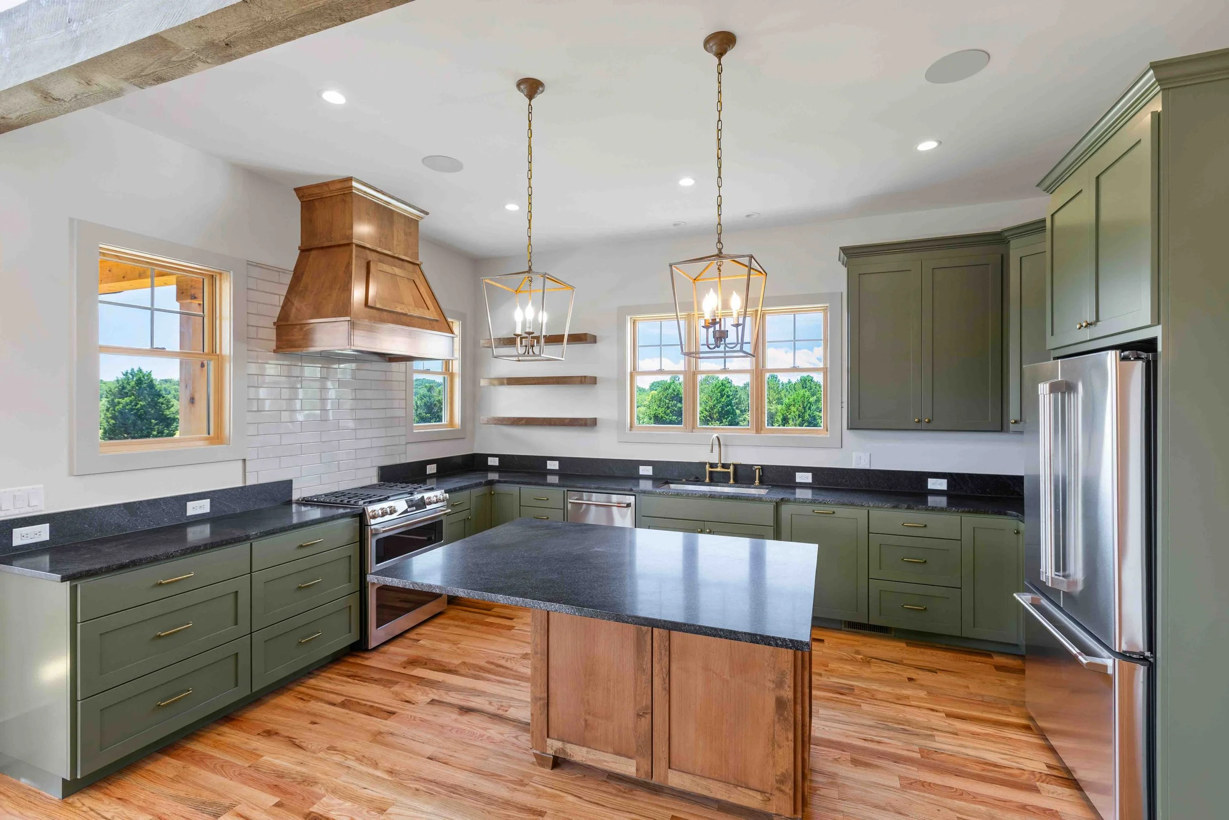 Modern kitchen with green cabinets, black countertops, wooden flooring, and a white brick backsplash. It features a kitchen island, stainless steel appliances, and two pendant lights hanging from the ceiling.