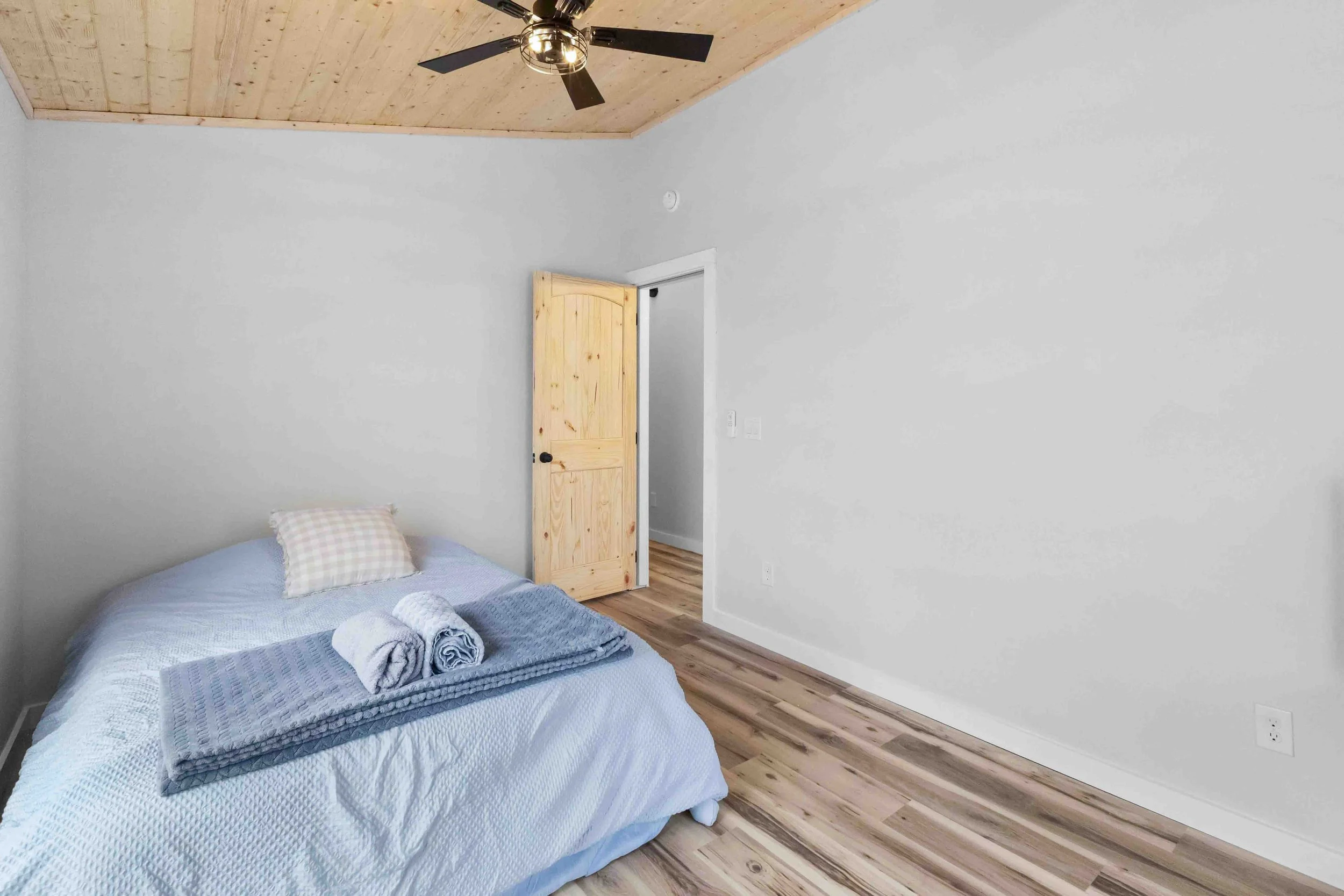 A bedroom with a wooden ceiling, white walls, a ceiling fan, a bed with white bedding and rolled towels, a plaid pillow, a wood door, and hardwood flooring.