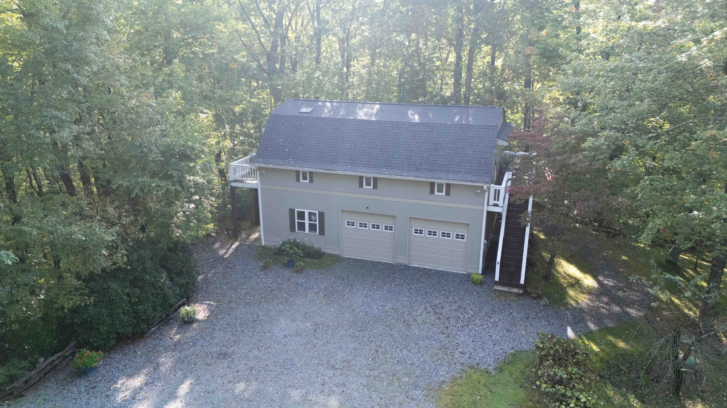 Aerial view of a two-story house with a gray exterior, three garage doors on the lower level, surrounded by trees and a gravel driveway.