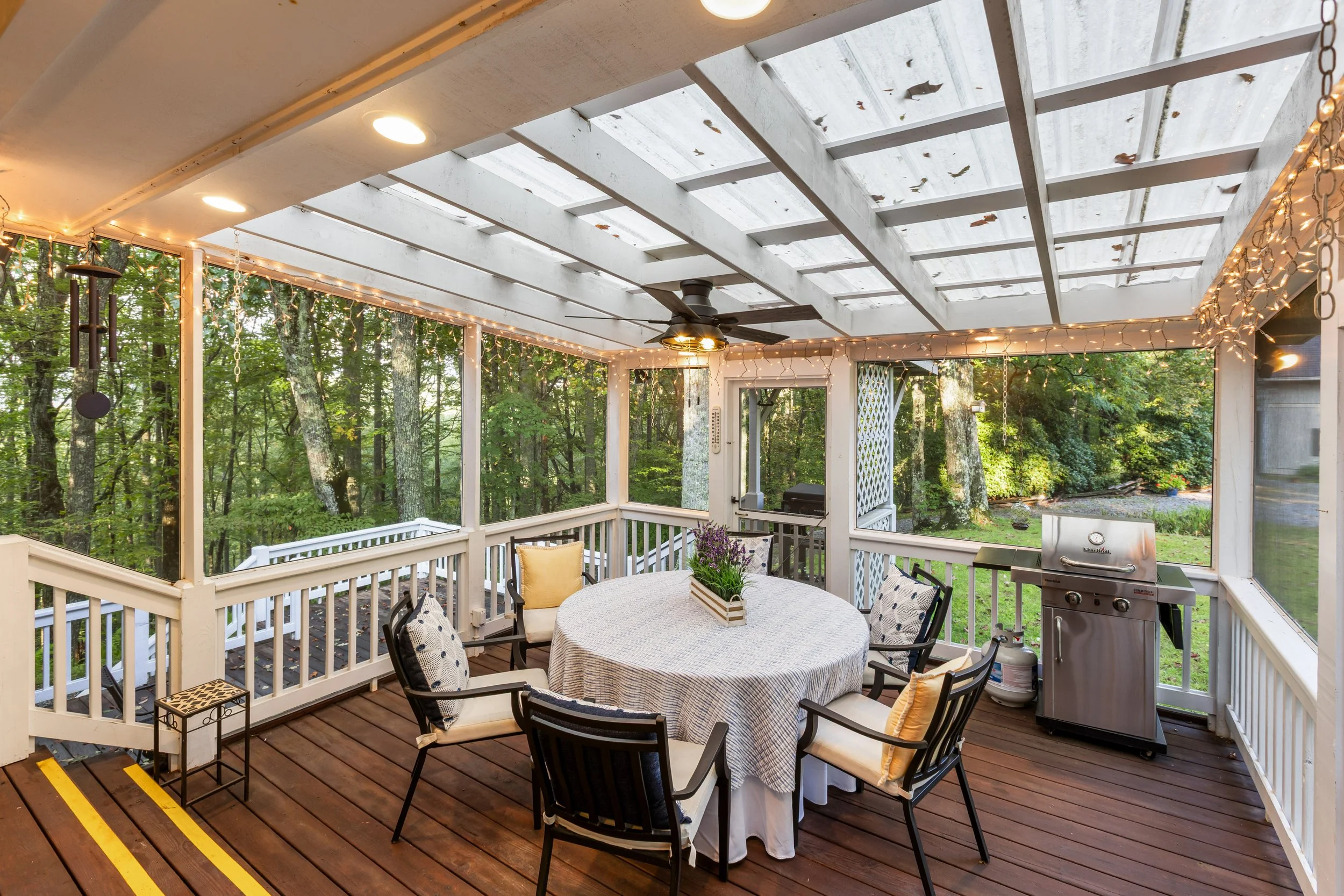 Covered porch with a round dining table, six chairs with cushions, and a potted plant centerpiece, surrounded by string lights and a grilling area, overlooking a wooded yard.