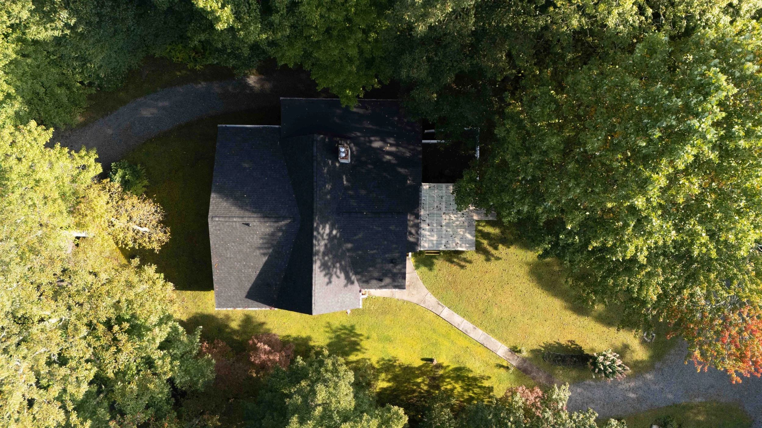 An aerial view of a house with a dark roof, surrounded by trees and a curved driveway with a grassy yard.