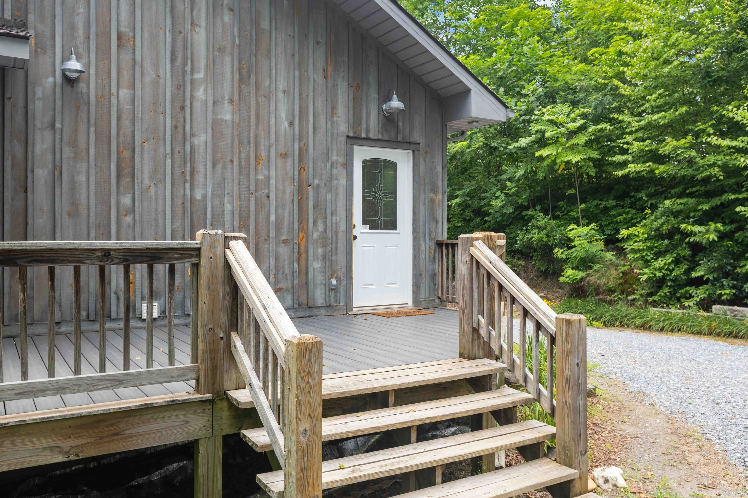 Wooden house with a small front porch, white door with decorative glass, and outdoor wall-mounted lights. Surrounded by green trees and a gravel pathway.