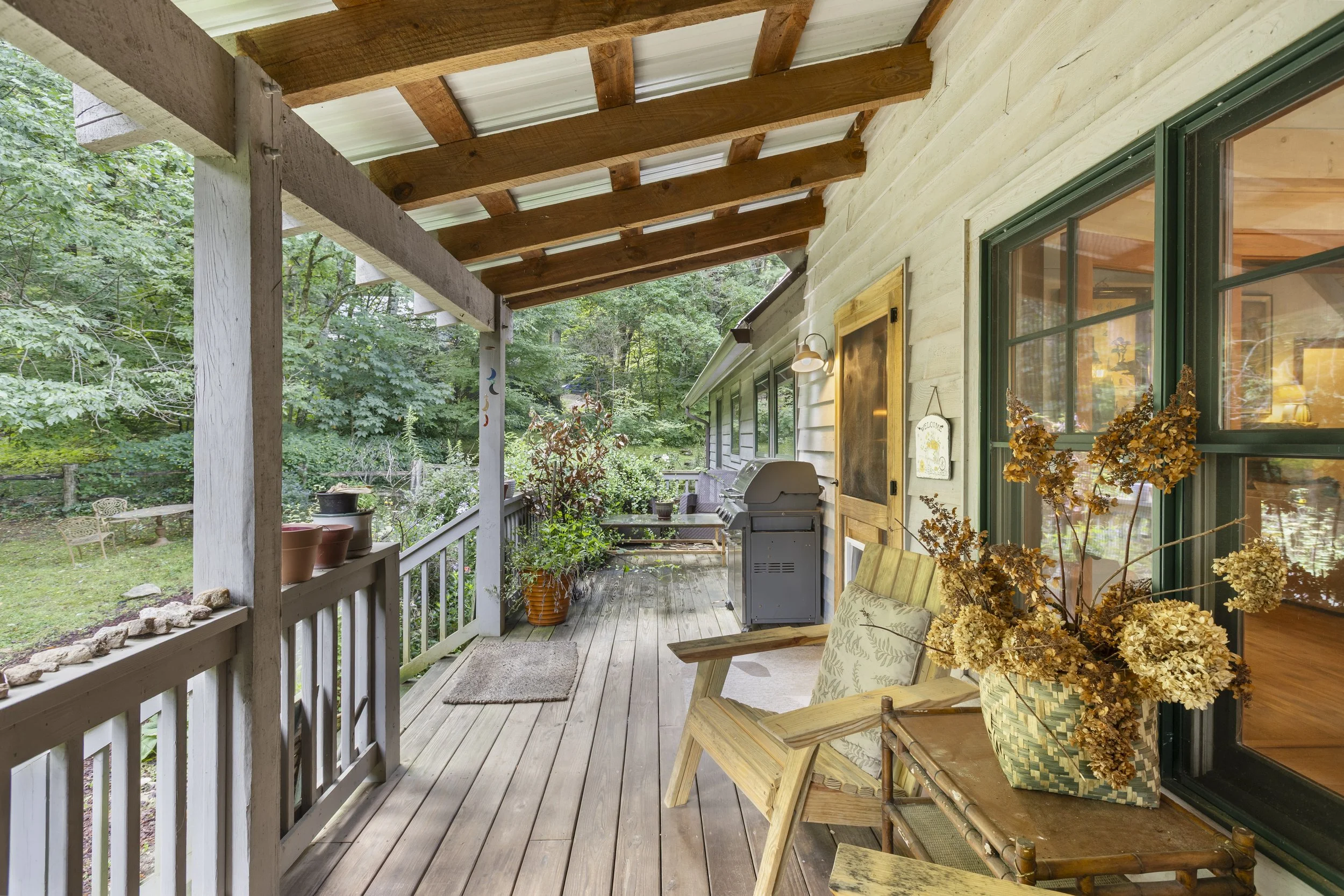 Image of a rustic porch with wooden flooring and railing, decorated with potted plants and dried flowers, with a view of lush green trees and a garden in the background.