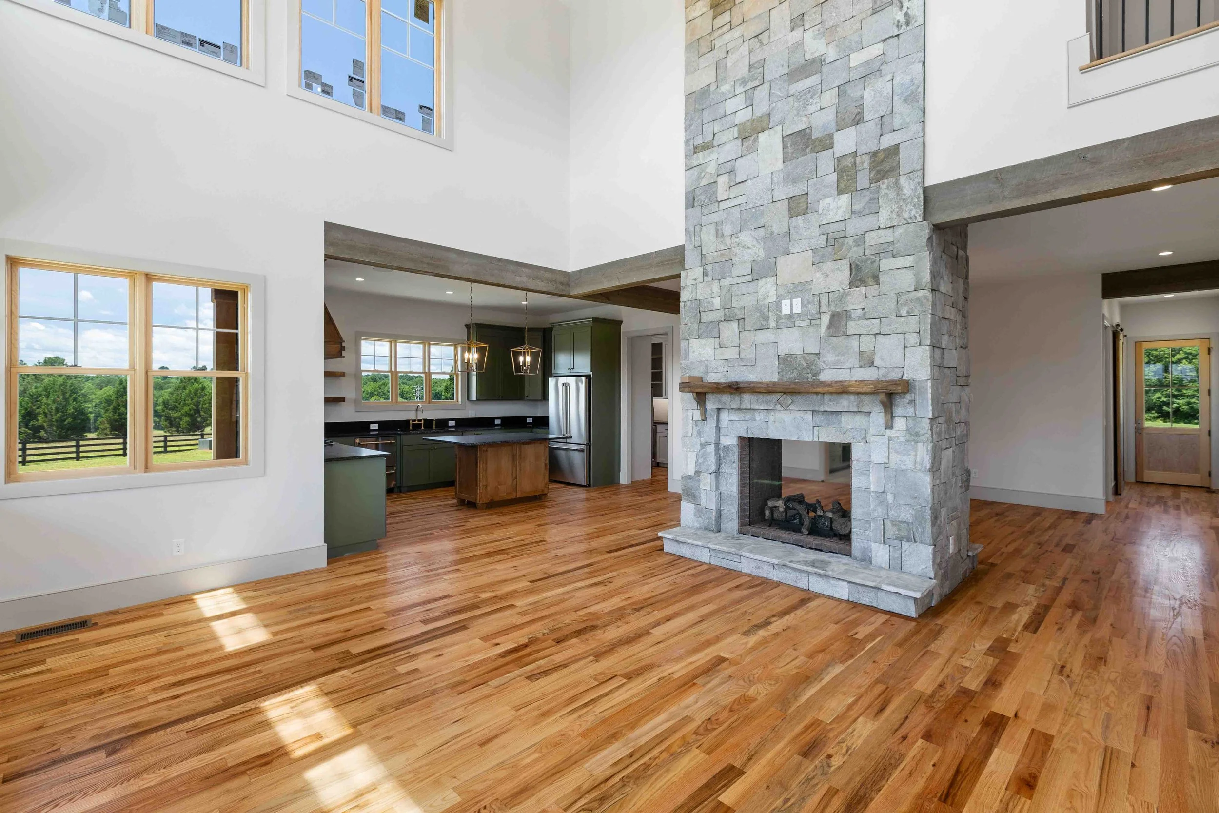 Living room with hardwood floors, stone fireplace, large windows, and an open kitchen with green cabinets and a kitchen island.