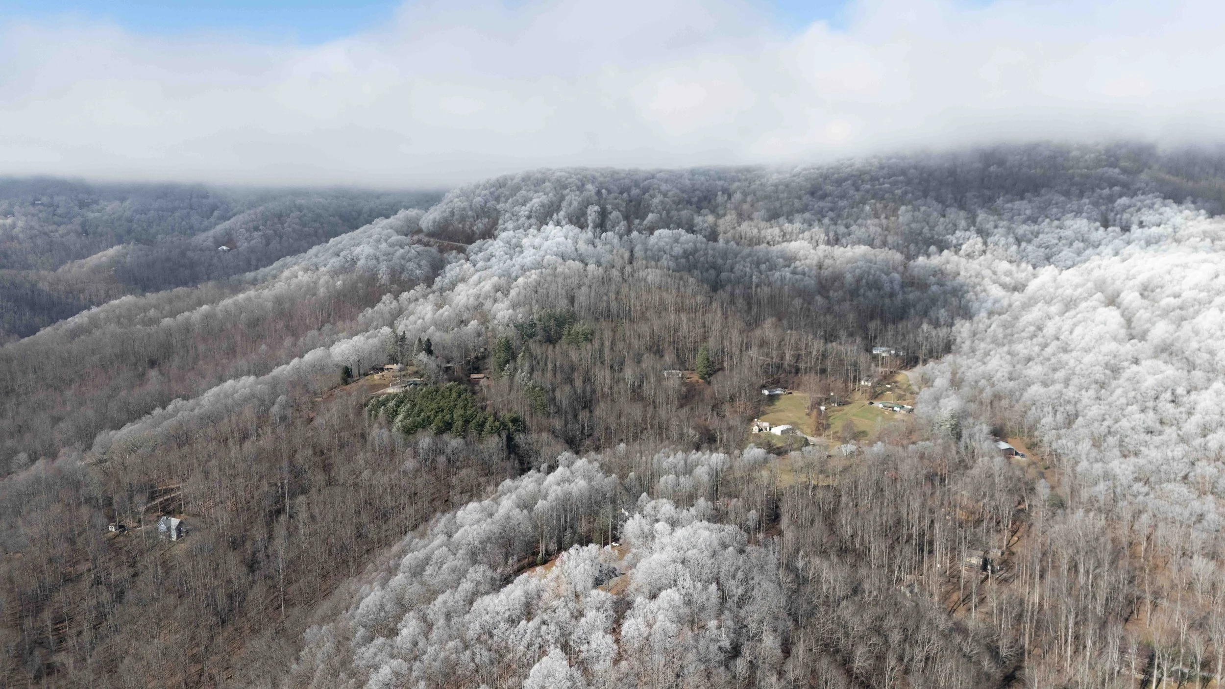 Aerial view of a mountain landscape with snow-covered trees and small houses scattered among the forest, with low clouds overhead.