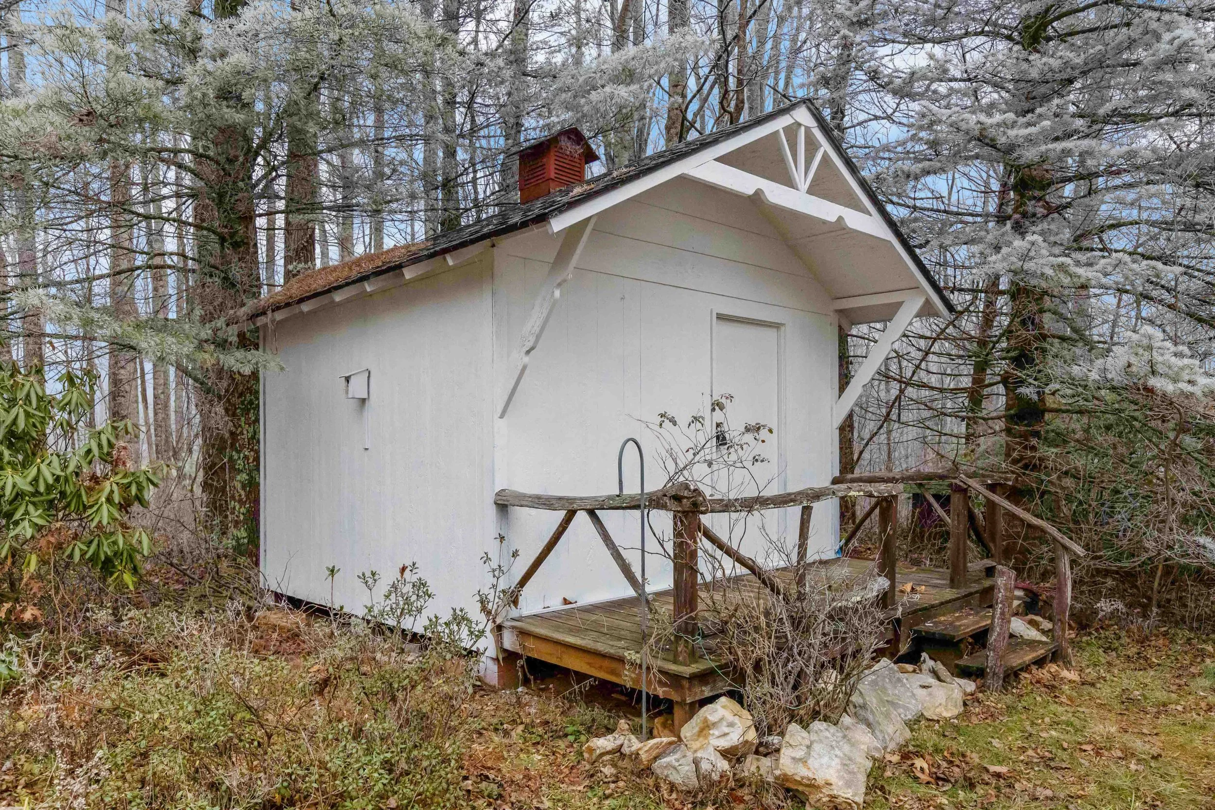 A small white shed in a wooded area with trees and bushes, featuring a ramp and a railing at the entrance, and a vent on the side.