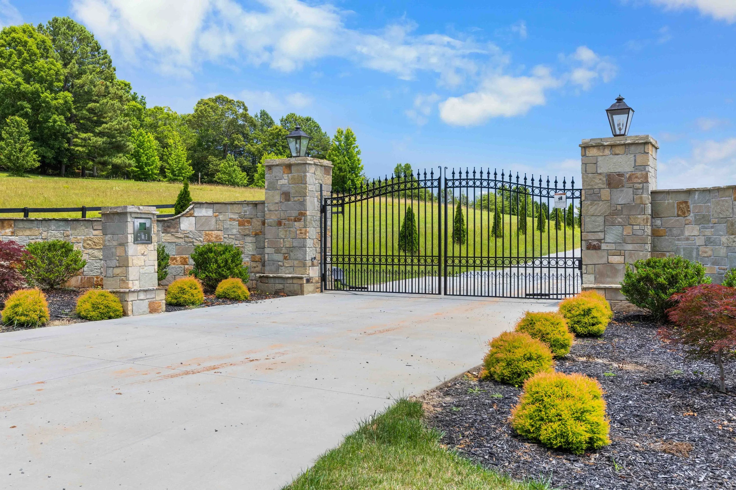Gated entrance with stone pillars and black wrought iron gates, surrounded by landscaped bushes and trees, leading to a scenic hilly area with green grass and a partly cloudy blue sky.