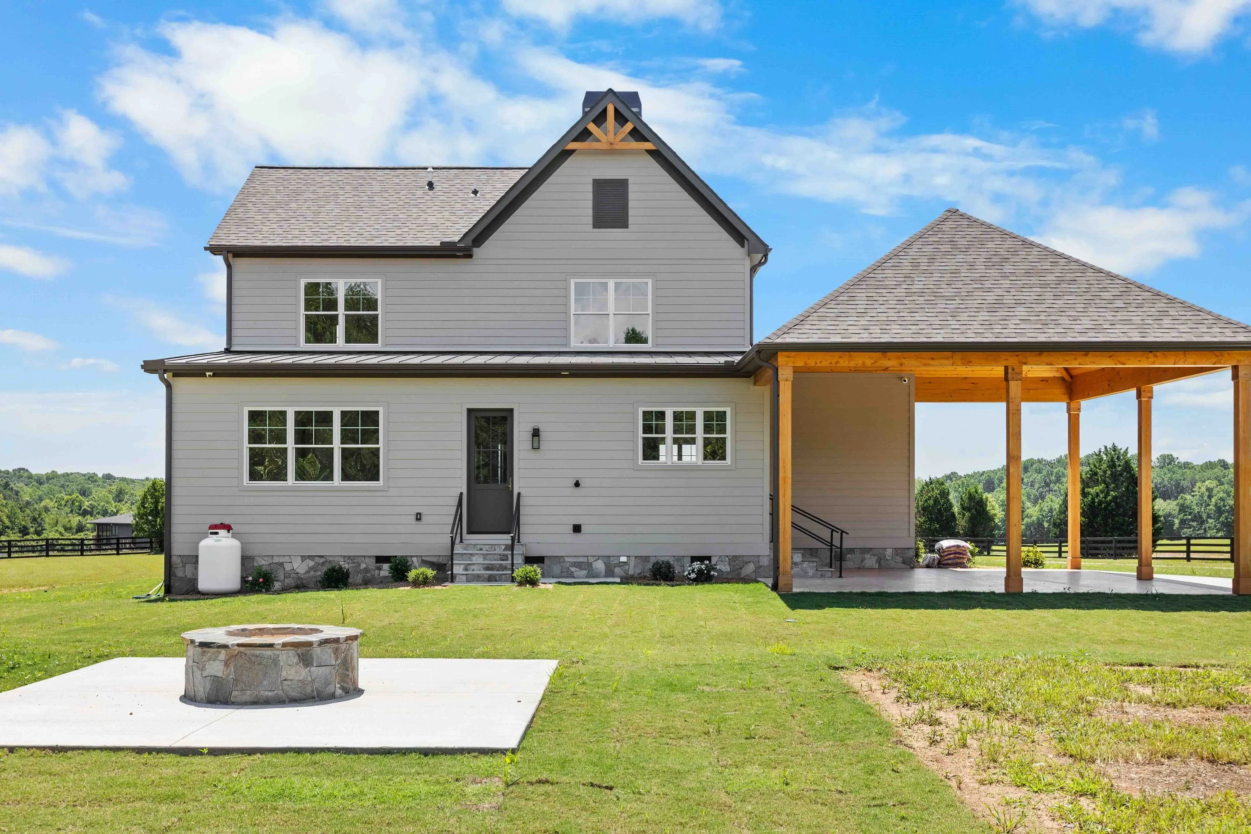 Front view of a two-story gray house with a small porch, a fire pit on the lawn, and a covered patio area with wooden posts, under a blue sky with clouds.