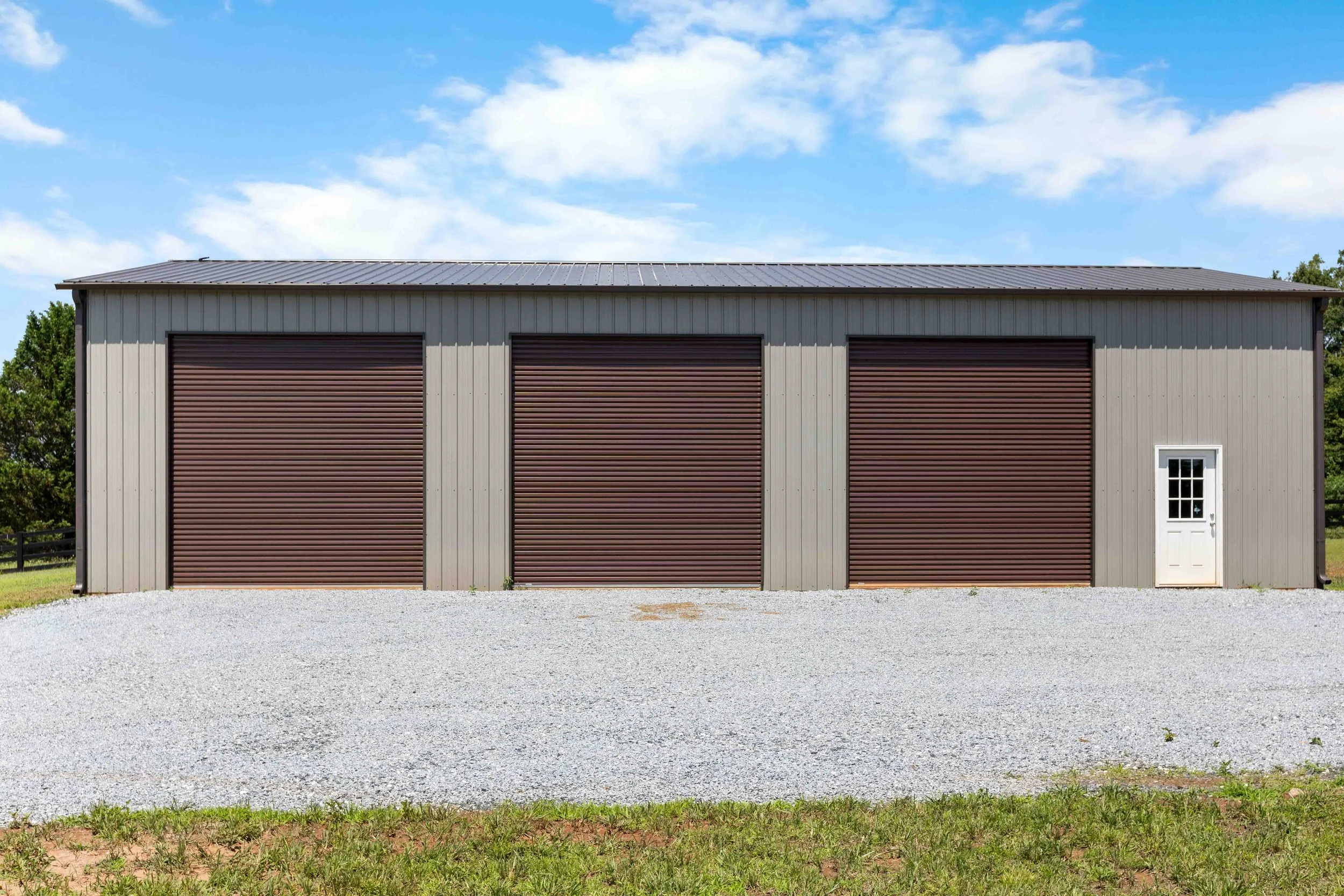A metal building with three closed brown roll-up garage doors and a white door with a window, situated on a gravel lot with grass and trees in the background under a blue sky with some clouds.
