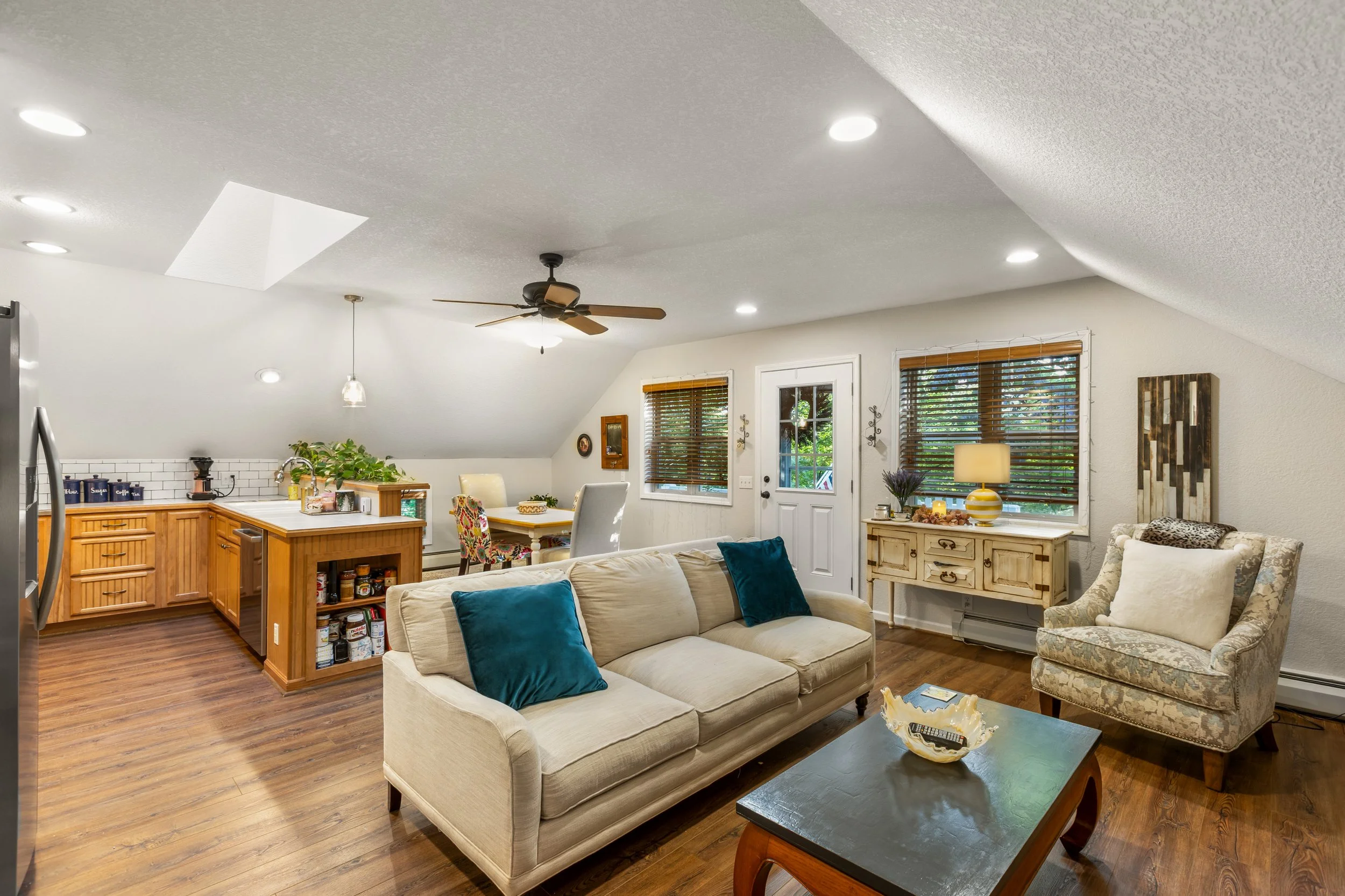 Living room with beige sofa, patterned armchair, wooden coffee table, and kitchen area in background with wooden cabinets and cutting board. Windows with blinds, hardwood flooring, white walls, and decorating items like lamp, plants, and wall art.