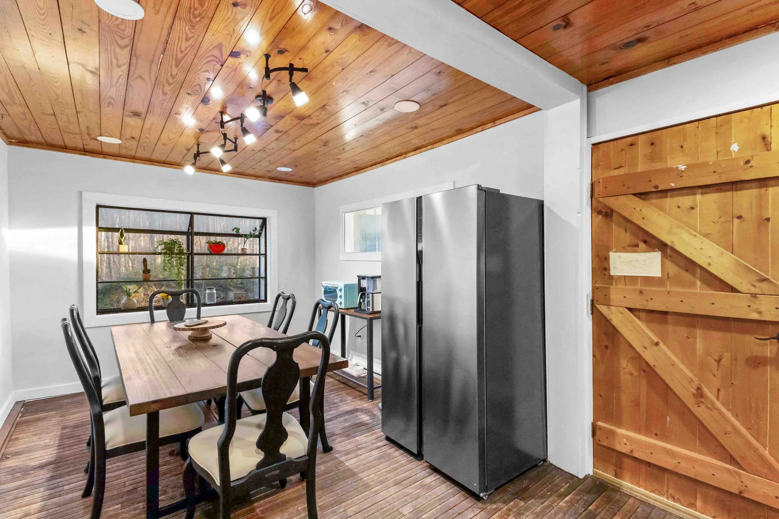 A dining room with a wooden ceiling, white walls, a large window with some plants, a wooden dining table with black and beige chairs, a black side table with a coffee maker and a small refrigerator, a metallic refrigerator, and a wooden door.