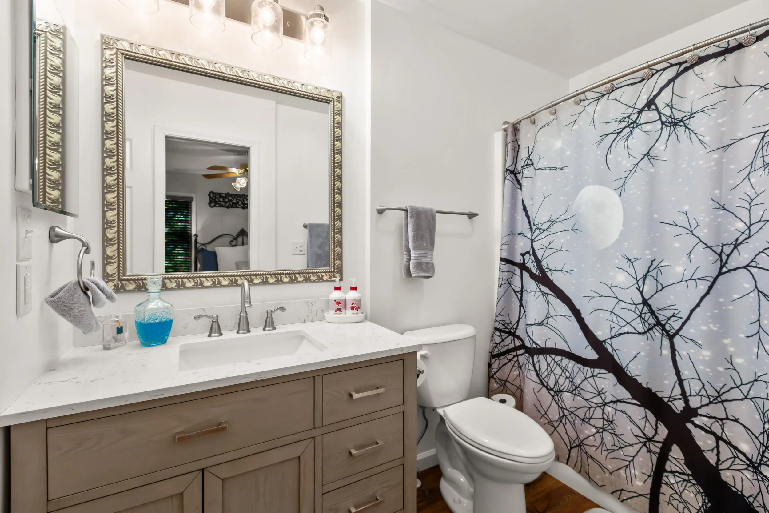 A bathroom with a white vanity top, a large decorated mirror, a faucet, a soap dispenser with blue liquid, two small bottles, a towel ring with a towel, a toilet, and a shower curtain featuring a nighttime scene with a full moon and tree branches.