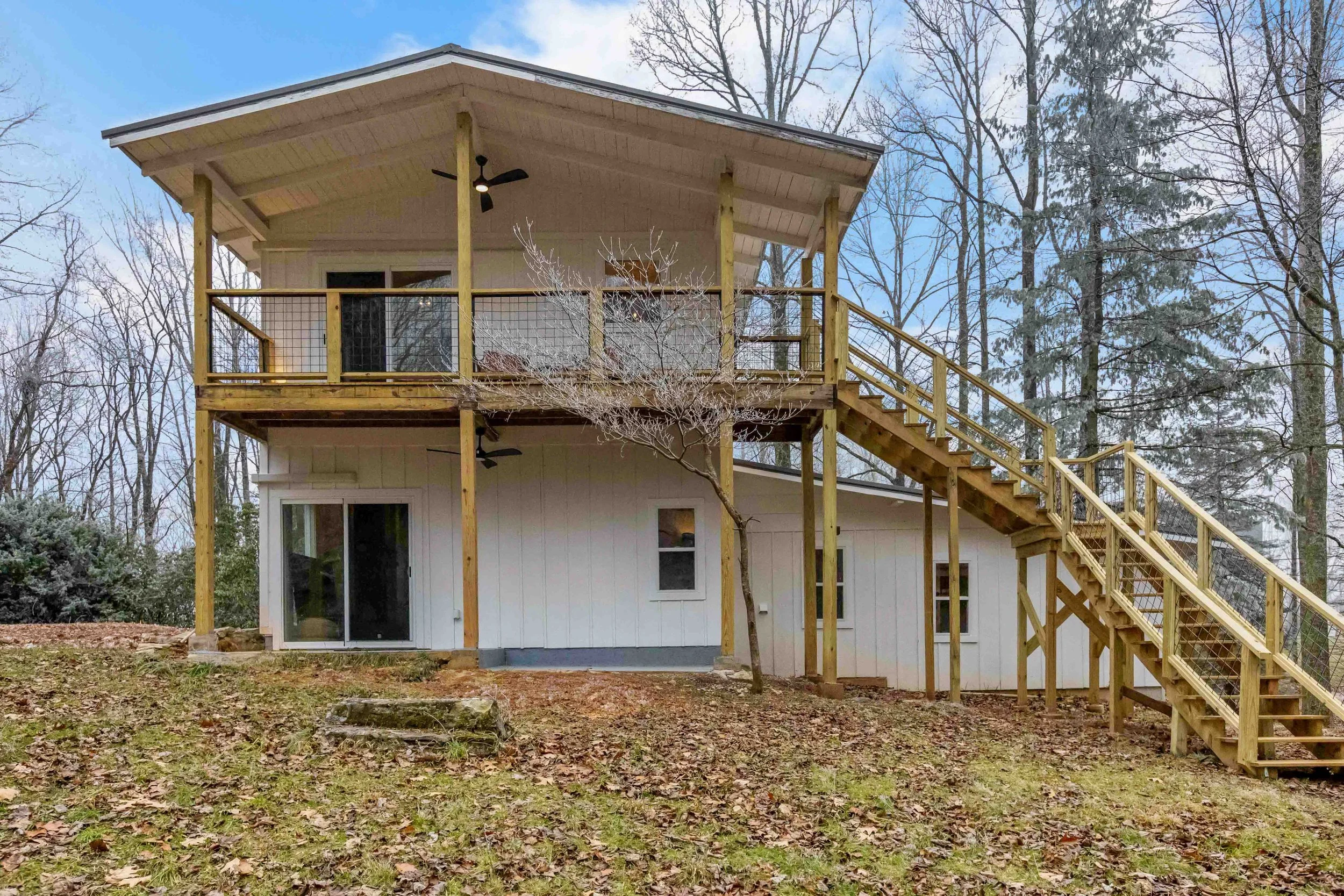 Two-story house with white siding and a large upper deck with a railing, accessed by wooden stairs, with trees and a partly cloudy sky in the background.