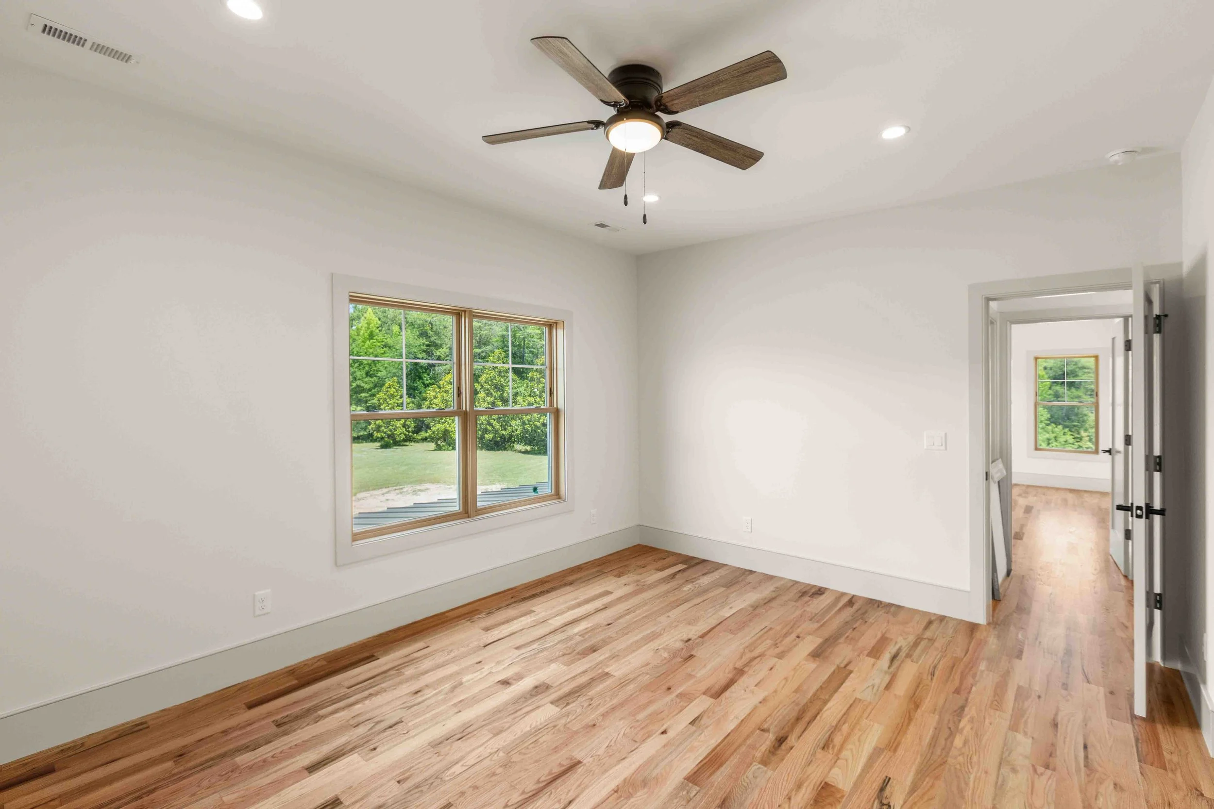 Empty room with white walls, a window letting in natural light, a wooden floor, a ceiling fan, and an open door leading to another room with a window.