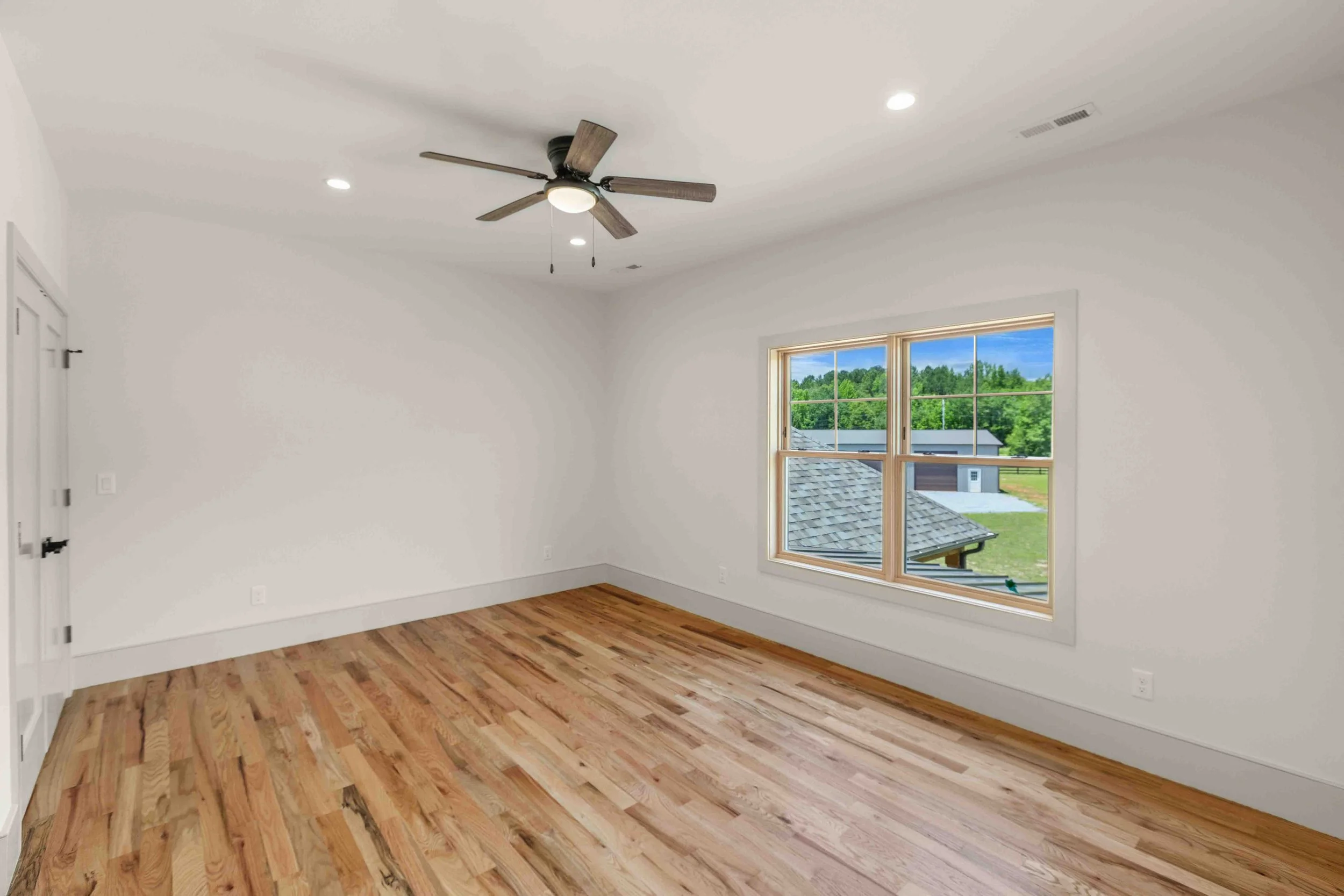 Empty room with white walls, hardwood floors, a ceiling fan, and a window showing a view of trees and a garage outside.