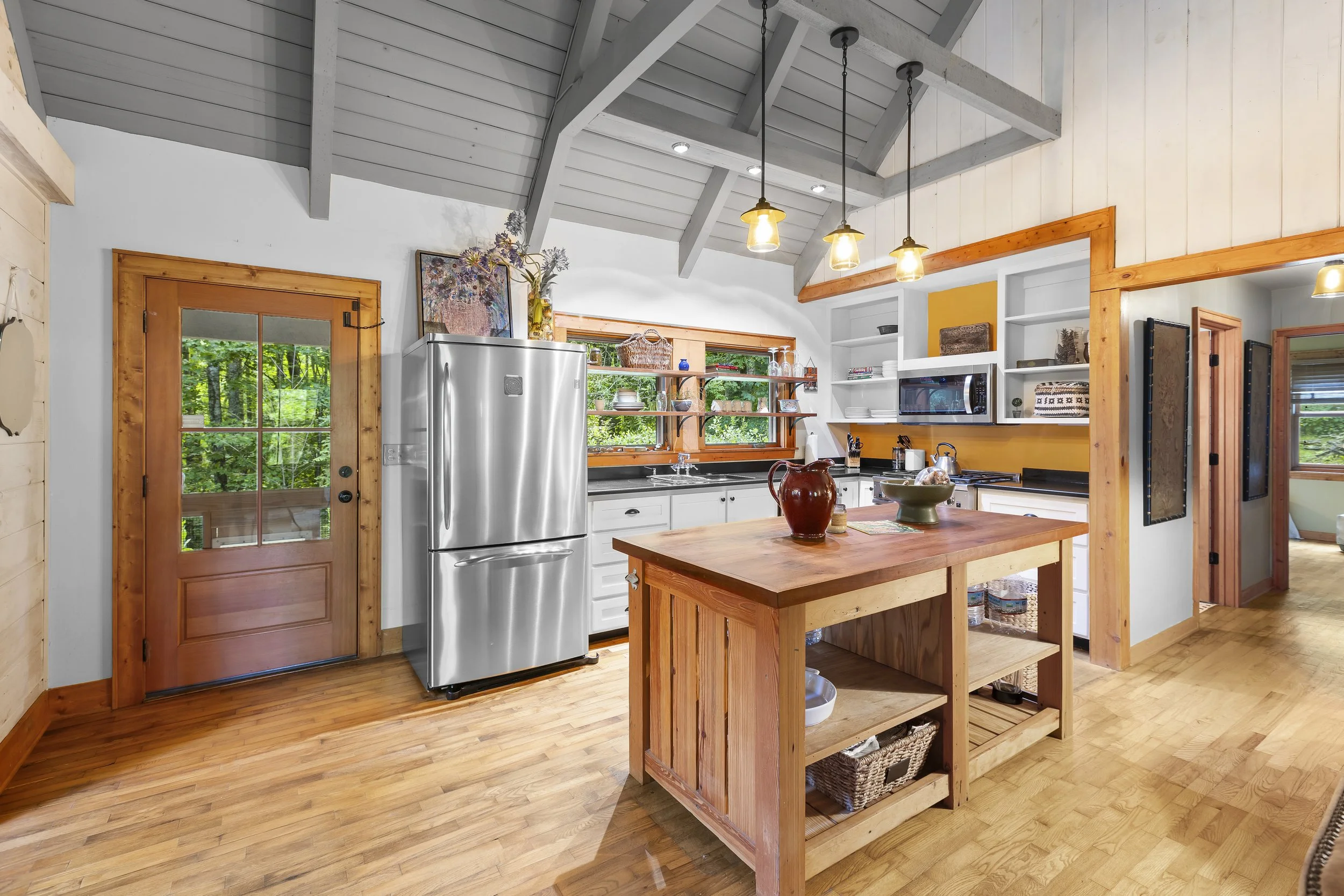 A kitchen with wooden floors, angled ceiling, white cabinets, a stainless steel refrigerator, wooden accents, open shelving, a window above the sink, and a kitchen island with bowls and decorative items.
