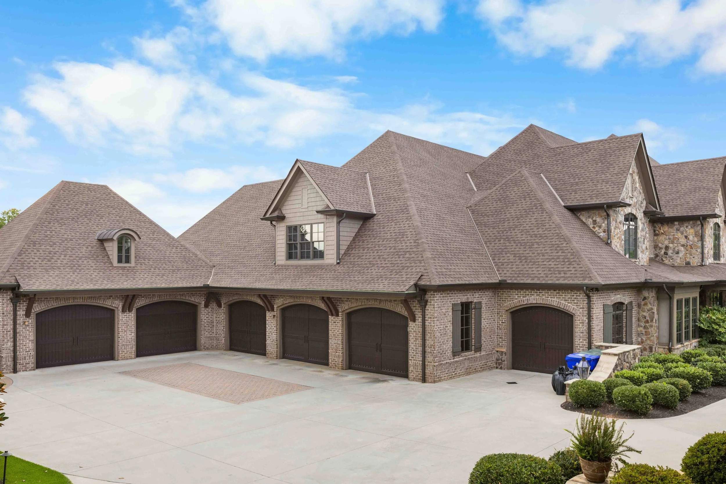 Modern large house with brick and stone exterior, multiple gabled roofs, and a three-car garage, surrounded by manicured shrubs and bushes, under a partly cloudy sky.