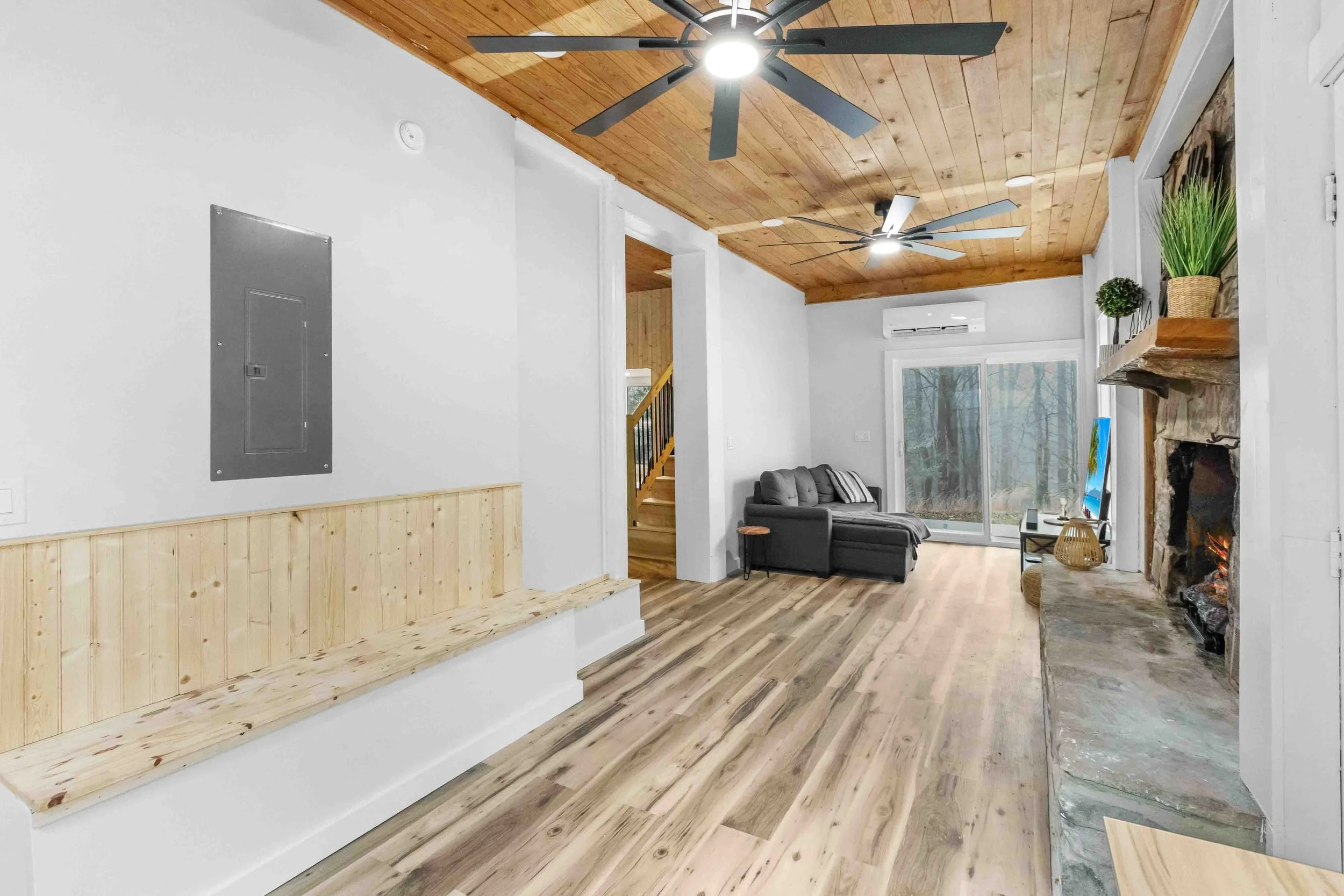 Living room with wooden ceiling, ceiling fans, gray sofa, fireplace with stone surround, sliding glass door to outside, wooden floor, potted plants, and minimal decor.