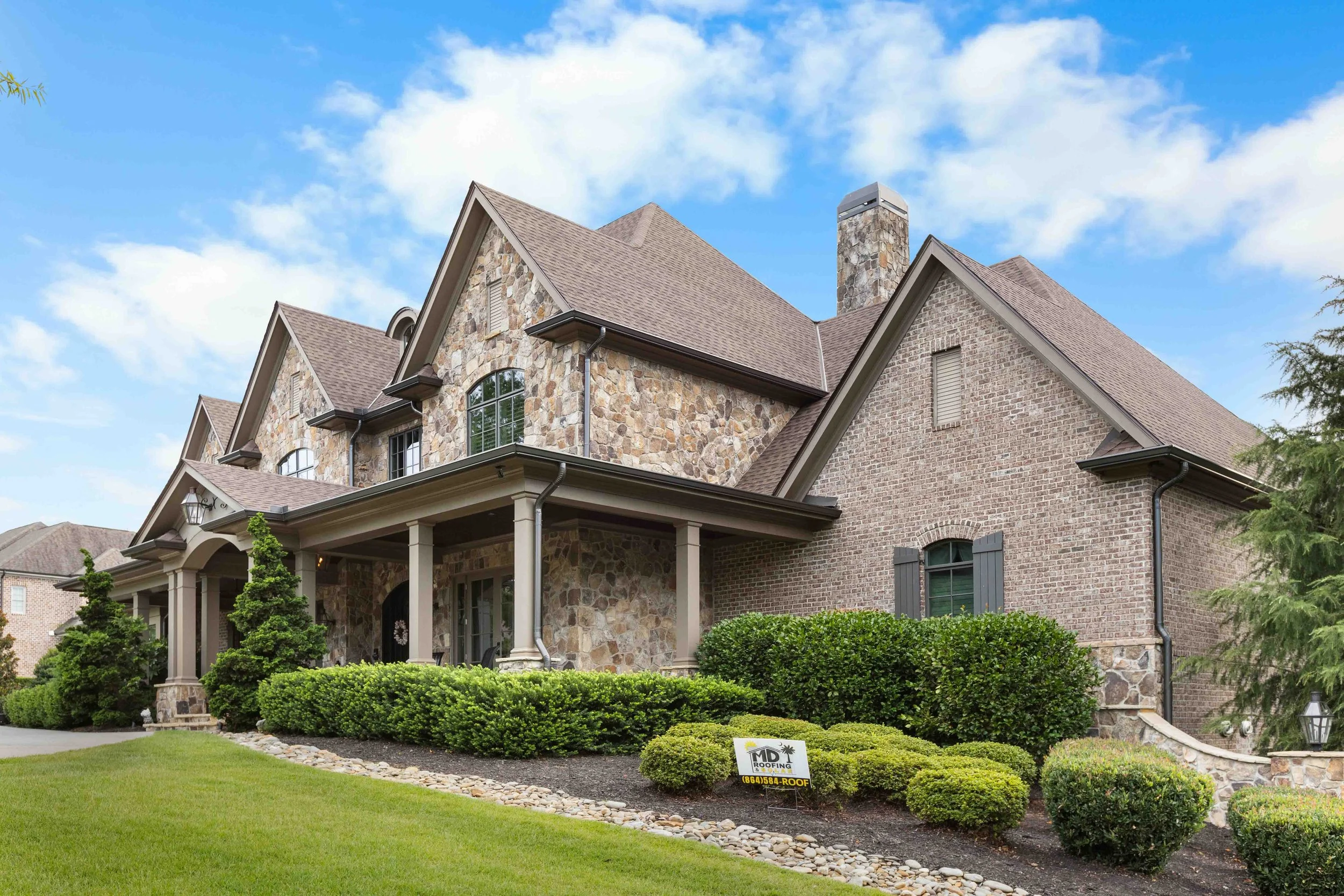Large house with stone and brick exterior, gabled roof, front porch, and well-maintained landscaping with shrubs and small trees.