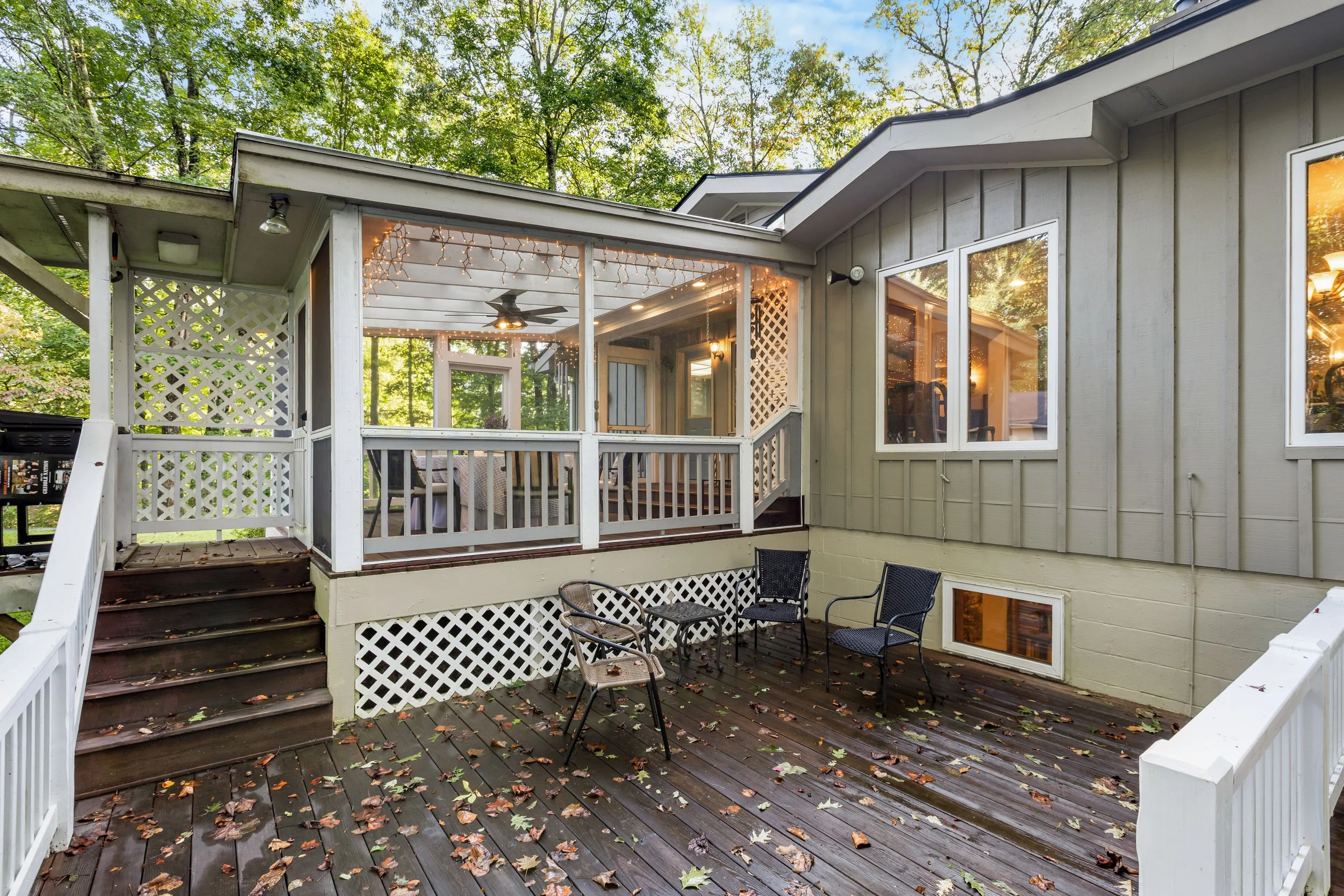 A wooden outdoor deck with four chairs, fallen leaves, and a screened-in porch with string lights, ceiling fan, and windows.