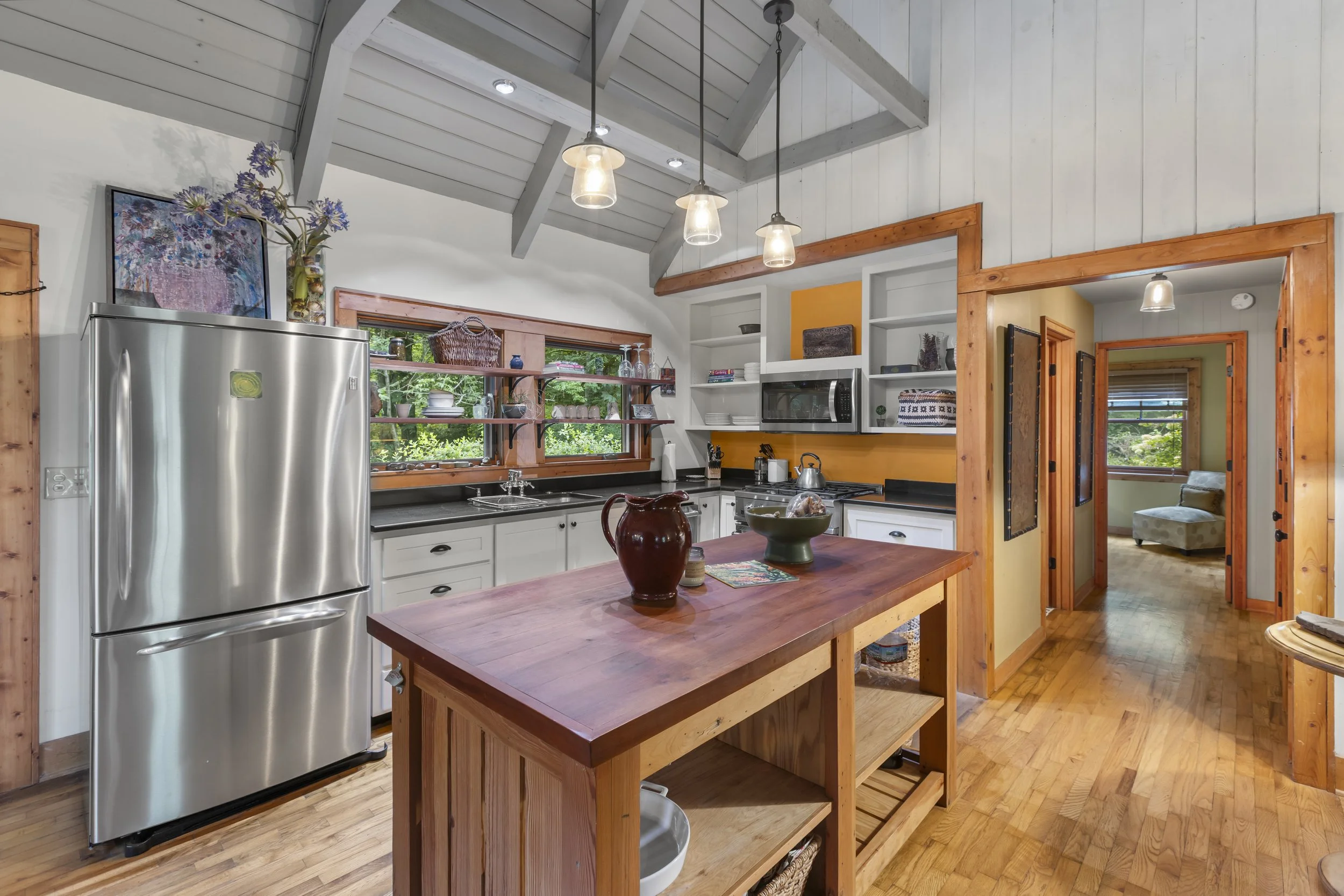 Kitchen with white cabinets, stainless steel refrigerator, wooden island, open shelves, black countertops, and a large window showing greenery outside.