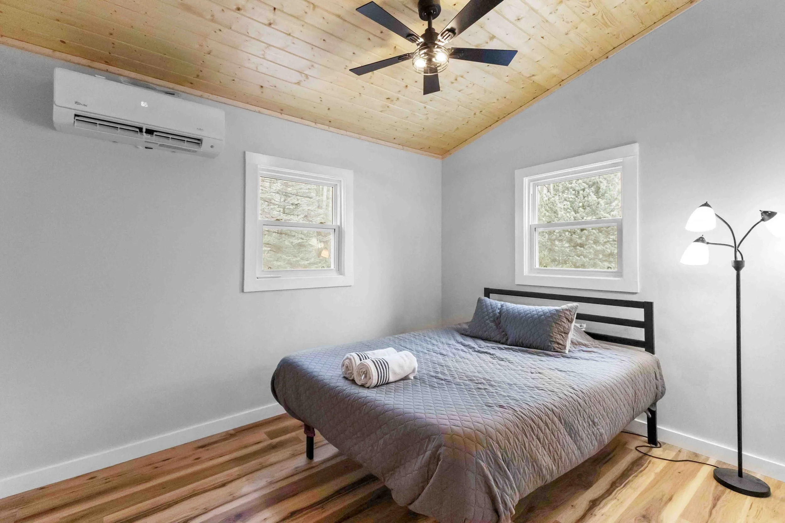 A bedroom with a wooden ceiling, white walls, a bed with gray bedding, two windows, a black floor lamp, and an air conditioning unit.