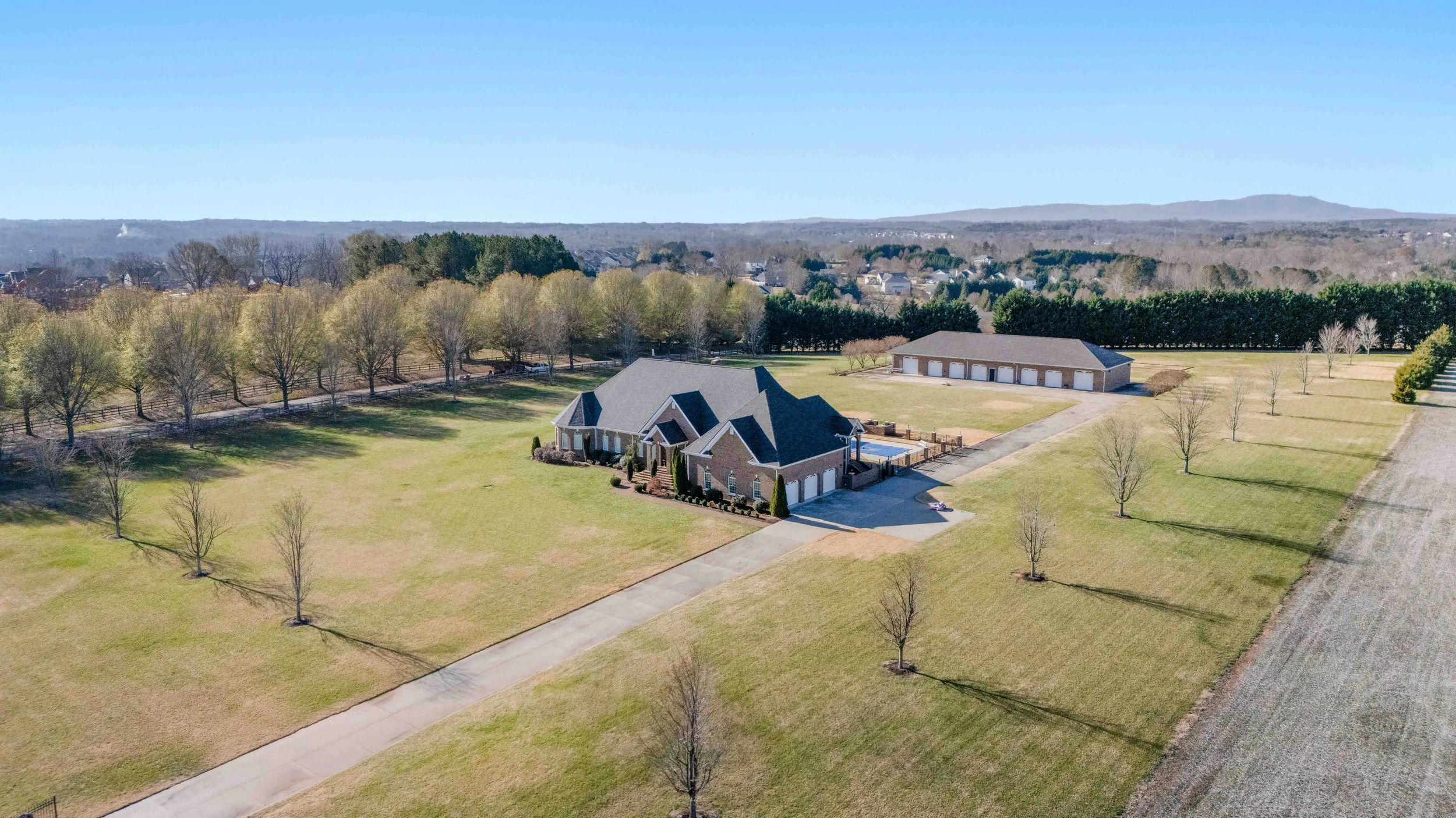 Aerial view of a large house with a spacious yard, driveway, and a detached garage or storage building, surrounded by trees and open land on a sunny day.