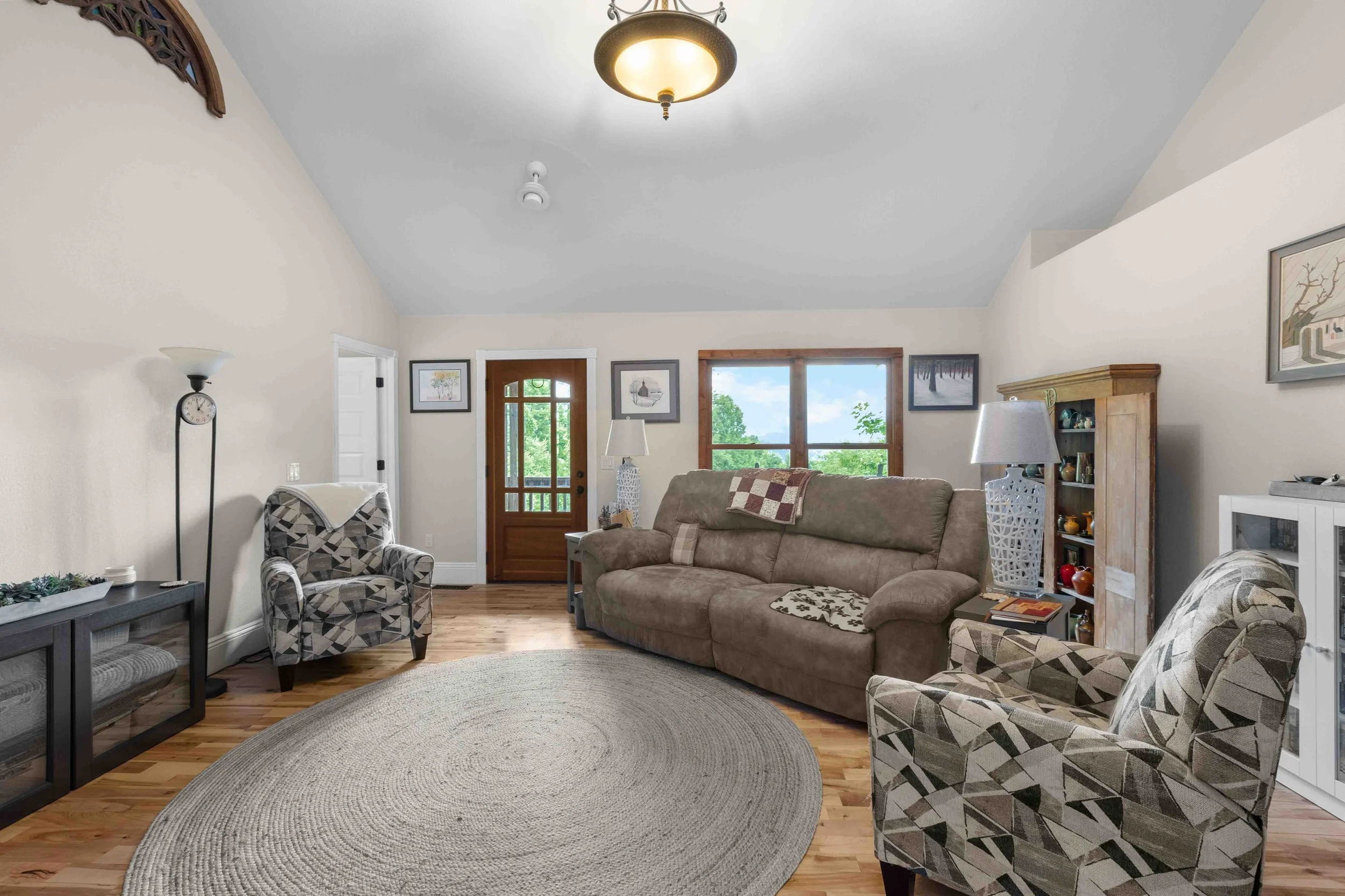 Living room with beige walls, hardwood floor, patterned armchairs, a beige sofa, a round braided rug, and wall art, with front door and windows showing greenery outside.